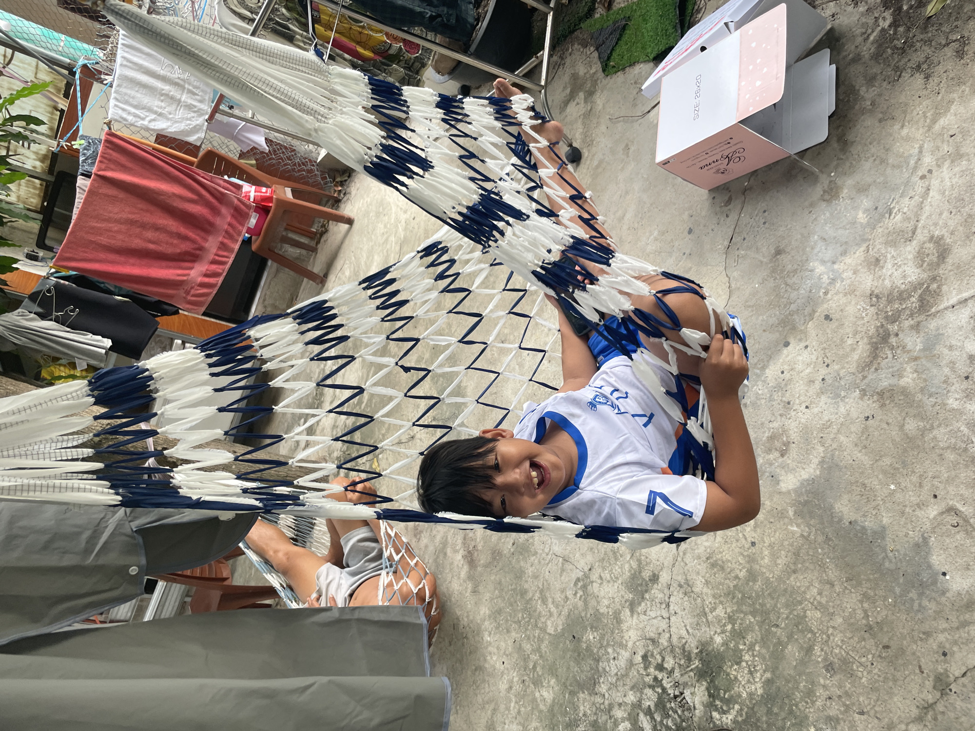 A young boy with dark hair and a white t-shirt with blue trim and a number 7 on the sleeve is laughing as he sits in a blue and white hammock. He is in a courtyard with a concrete floor. In the background, there is laundry hanging on a line, a chair, and a metal fence. A portion of another person's legs and feet are visible in the upper left of the image. To the right of the boy, there is a pink and white cardboard box with text that reads "SIZE: 25x20". The scene appears to be outdoors, possibly in a residential area.