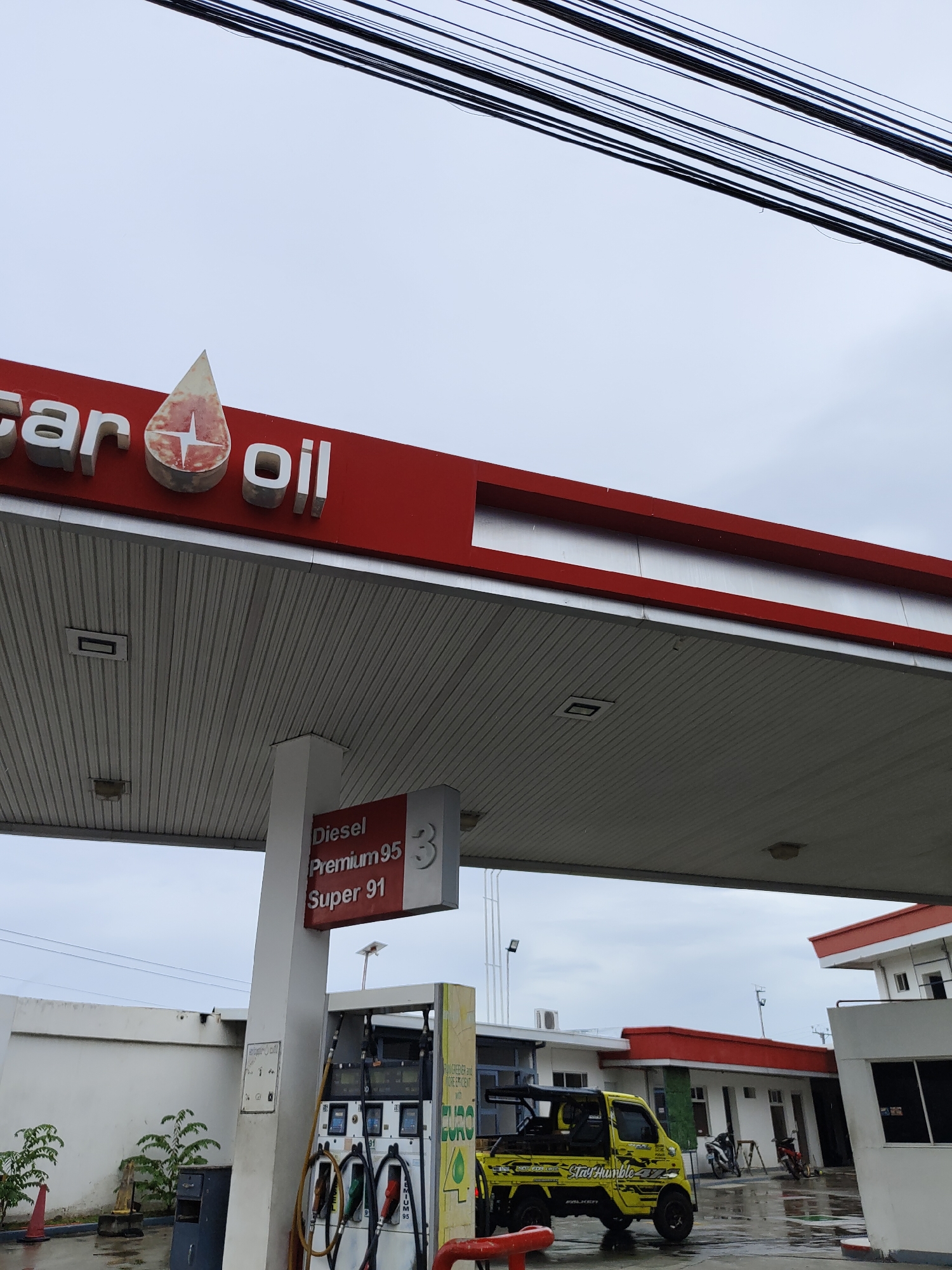 This image depicts a street-level view of a gas station in Davao City, Philippines, under an overcast sky. The foreground is dominated by the canopy of the gas station, featuring a red and silver structure with corrugated metal underside. Prominently displayed on a white pillar is a sign indicating fuel types: "Diesel," "Premium 95," and "Super 91."
Below the canopy, a yellow mini truck with black accents and the phrase "Stay Humble" is parked. It has a distinctive aftermarket roof rack and decals. To the left of the truck, a row of fuel pumps is visible, each with a hose. The ground is wet, suggesting recent rain.
In the background, a white building with red trim is visible, possibly part of the gas station's convenience store or office. Another building, also with red trim, is seen further back. A few motorcycles are parked near the building. A white wall runs along the left side of the image, with some sparse greenery and a red traffic cone. Overhead, a dense network of black electrical wires crisscrosses the sky. The overall scene suggests a typical day at a local gas station in the Philippines.