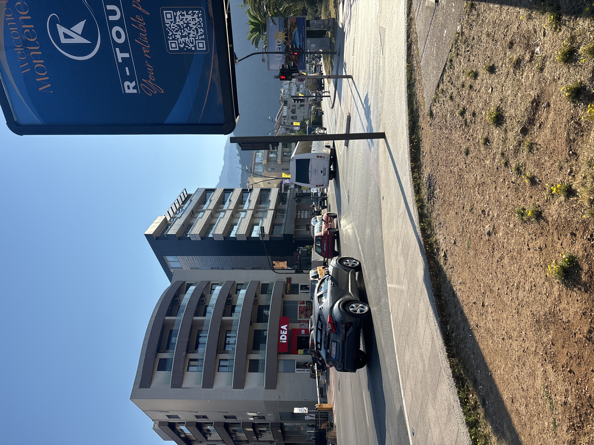 The image depicts a street scene in Budva, Montenegro, on a clear, sunny day. In the foreground, a dark gray car is driving on the right side of the road. Behind it, a red car and a smaller orange vehicle are also visible. The road is lined with a sidewalk and a patch of dry, brown earth with scattered yellow wildflowers.
In the middle ground, several modern buildings with balconies and glass railings dominate the urban landscape. One building features a prominent red "iDEA" sign. Across the street, traffic lights are visible, with one currently showing red. A white tour bus is turning at an intersection.
In the background, more buildings and mountains are silhouetted against the bright blue sky. A large blue advertisement billboard with text and a QR code is positioned on the right side of the frame, displaying the words "Welcome to Montenegro" and "R-TOUR Your reliable p". Palm trees are also present in the background, suggesting a coastal or southern European location. The overall impression is of a bustling urban environment in a warm climate.