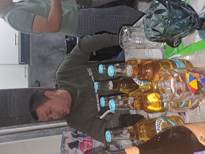 A casual indoor gathering shows a man in a green sweater near multiple bottles of "Pulco" beer, with light blue caps. There are several glasses present, including one with a beach umbrella design and a large, decorative glass container. Another person is visible in a green shirt and gray pants. The setting appears to be a kitchen or dining area with white cabinets and a gray countertop.