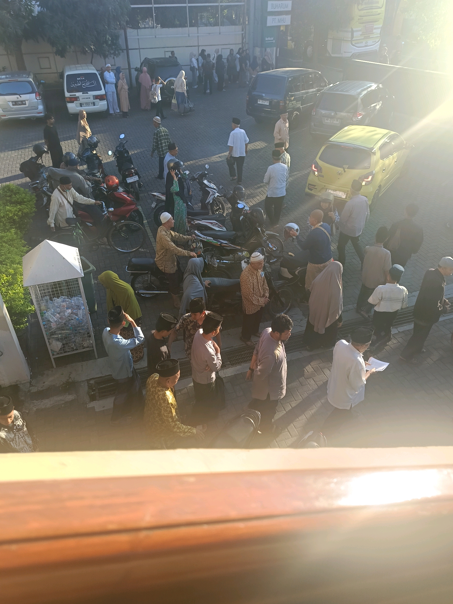 The image shows an outdoor scene with many people and vehicles, suggesting a public gathering or event. The bright sunlight indicates it is daytime, and the presence of vehicles like cars and motorbikes points to a location with accessibility. The architectural style of the buildings in the background, particularly the details of the windows and the overall structure, could help in identifying the specific location.
The people in the image are diverse, with some wearing traditional clothing, which is common in Indonesia. The presence of women in hijabs and men in various attire, including some in white shirts and caps, suggests a cultural or religious context. The interactions between people, such as conversations or walking together, provide clues about the nature of the event.
The environment includes a paved ground, possibly a parking lot or a public square, with a white structure that appears to be a recycling bin filled with plastic bottles. The foliage of trees on the left side of the image adds a natural element to the urban setting. The bright sun glare in the upper right corner of the image suggests the time of day and the direction of the sun.
Considering the provided context, this image is likely taken in the city of Yogyakarta, Indonesia, a city known for its rich cultural heritage and strong Islamic influence. The presence of specific vehicle license plates, if visible and decipherable, could potentially confirm the location. Without further information or visual cues that specifically point to Yogyakarta, it's a reasonable inference based on the typical scenes observed in Indonesian cities, particularly those with a significant Muslim population. However, for a forensic description, this inference would be noted as a hypothesis rather than a confirmed fact.