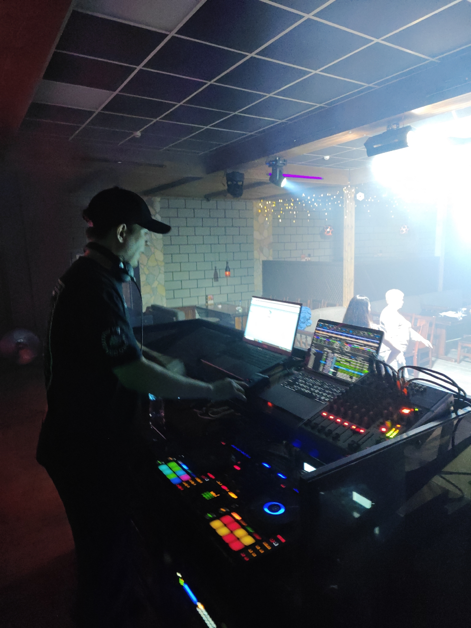 A DJ is at work in a dark nightclub or lounge in Chaikovsky, Russia. He is wearing a dark shirt and a baseball cap, and has headphones around his neck. In front of him is a complex array of DJ equipment including two laptops, a mixing console, and a MIDI controller. Behind him, patrons are seated at tables. The room is illuminated with a combination of overhead lighting and stage lights. The walls are decorated with brick-like patterns and string lights.