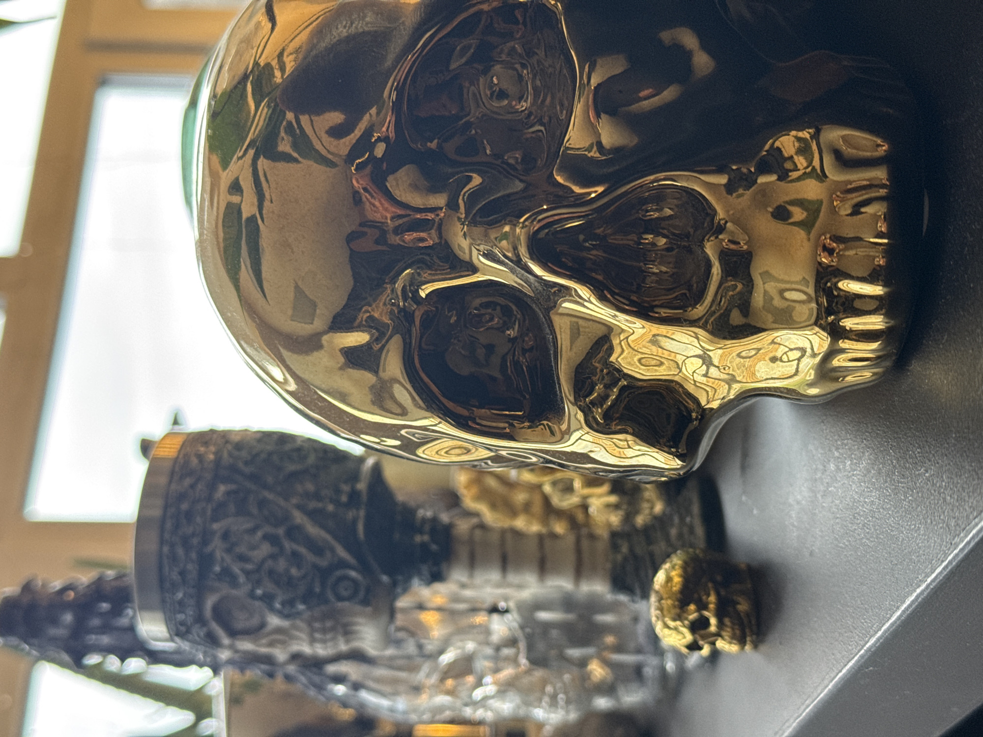 The image shows a collection of decorative skulls. The largest skull is gold and highly reflective, positioned prominently in the foreground. Behind it, there is another decorative skull with more intricate, darker detailing. In the background, partially obscured, is a smaller gold skull. All of the skulls are placed on a dark-colored surface, likely a shelf. Light from a window illuminates the scene, creating reflections on the gold skull. There is no indication that the photograph was taken in Krasnoyarsk. No people, actions or interactions are present in the scene.