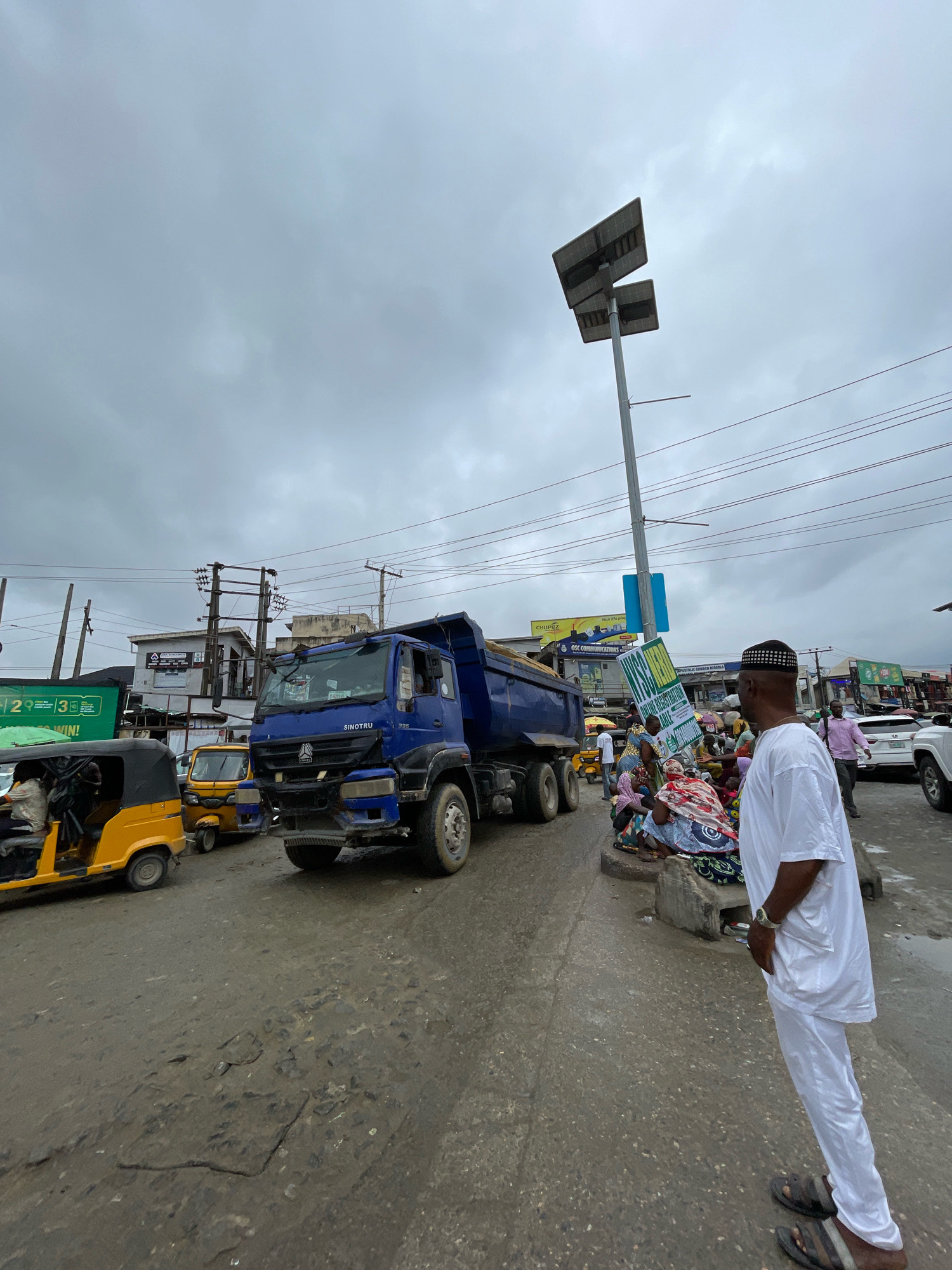 In Ajah, Nigeria, a cloudy day looms over a bustling street scene. A large blue dump truck, emblazoned with "SINOTRUK," dominates the foreground, its massive tires resting on a wet, pothole-ridden asphalt road. To the left, a yellow auto-rickshaw, a common mode of transport in the region, carries passengers. Further down the street, more auto-rickshaws are visible, along with a white SUV.
A group of people, dressed in colorful attire and traditional clothing, are gathered near a concrete ledge. Some are seated, while others stand, interacting and conversing. Among them, a man in a white traditional garment and a patterned cap stands facing away from the camera, his hands in his pockets, observing the scene. A lamppost with solar panels stands tall against the overcast sky, an indicator of modern infrastructure amidst the urban activity.
The environment is characteristic of a busy Nigerian city: a mix of commercial buildings, advertisements, and overhead power lines. The overall impression is one of vibrant street life and a community going about its daily business under a somber sky.