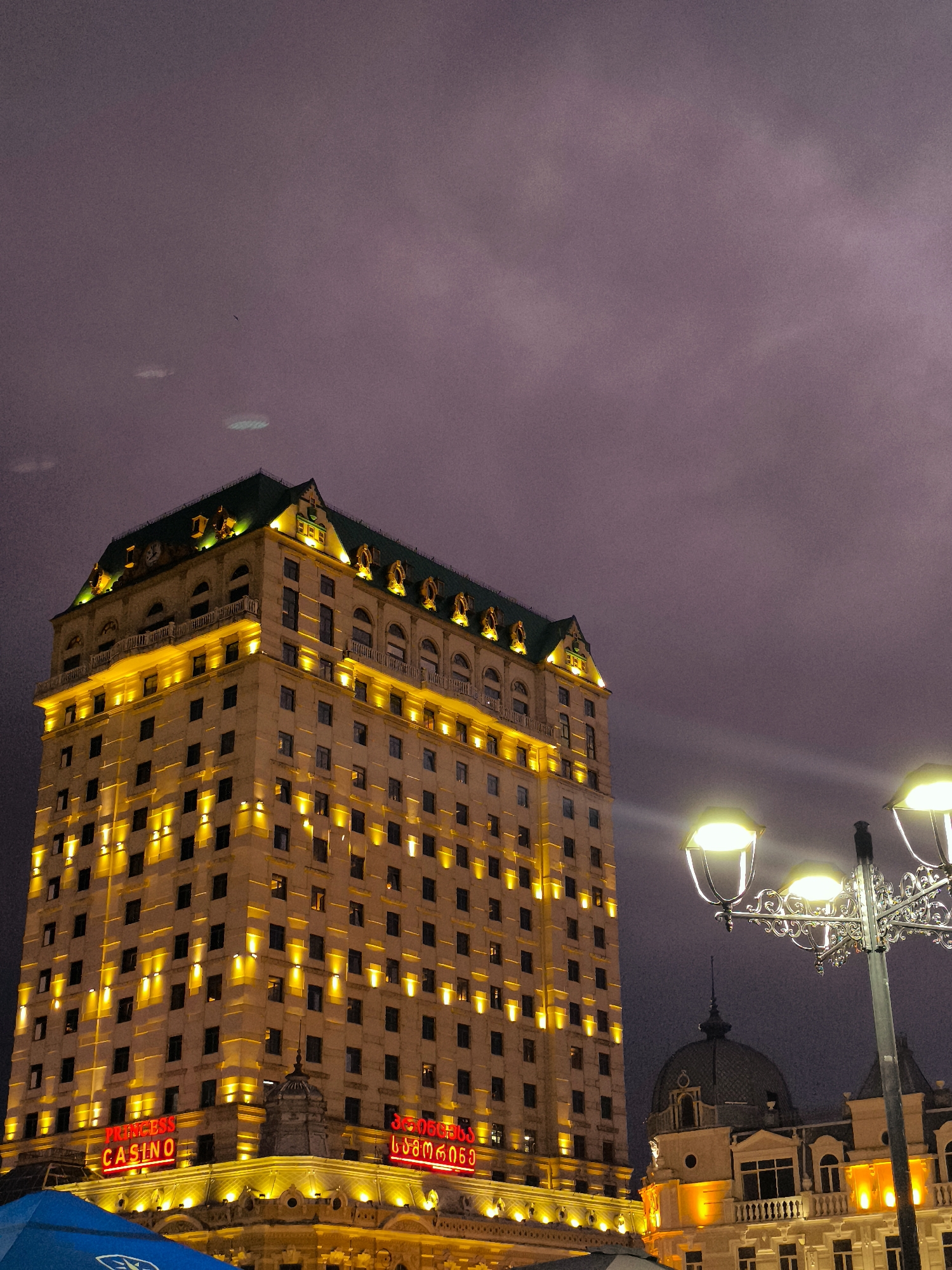 The image shows the illuminated exterior of the Sheraton Batumi Hotel, a tall, light-colored building with a green, mansard-style roof. The hotel is lit with numerous yellow lights, highlighting its many windows. Red neon signs on the lower floors read "PRINCESS CASINO" and also include text in Georgian script. To the right of the hotel, a decorative lamppost with three lights illuminates the foreground. In the background, the sky is a dusky purple with some light clouds. Adjacent to the Sheraton, another building with a dark dome and illuminated windows is visible. The scene appears to be in the city of Batumi, Georgia, at dusk or night. There are no people clearly visible in the image.
