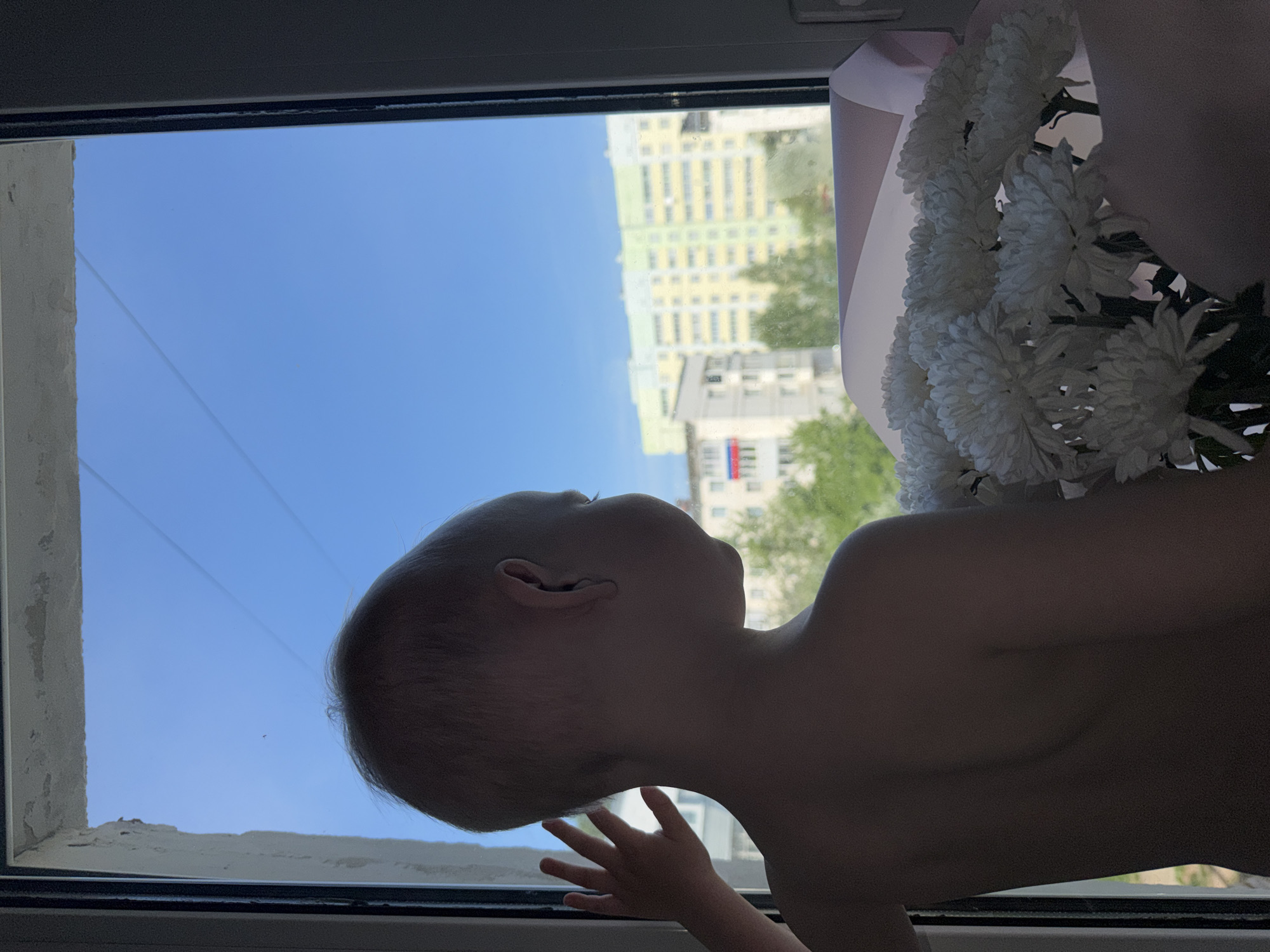 A young child stands topless at a window, peering outside. Their hand is placed against the lower window pane. A bouquet of white flowers, wrapped in pink paper, sits next to the window. Outside the window is a view of a multi-story building with a Russian flag hanging from a balcony, and a clear blue sky. The setting appears to be Chaikovsky, Russia.