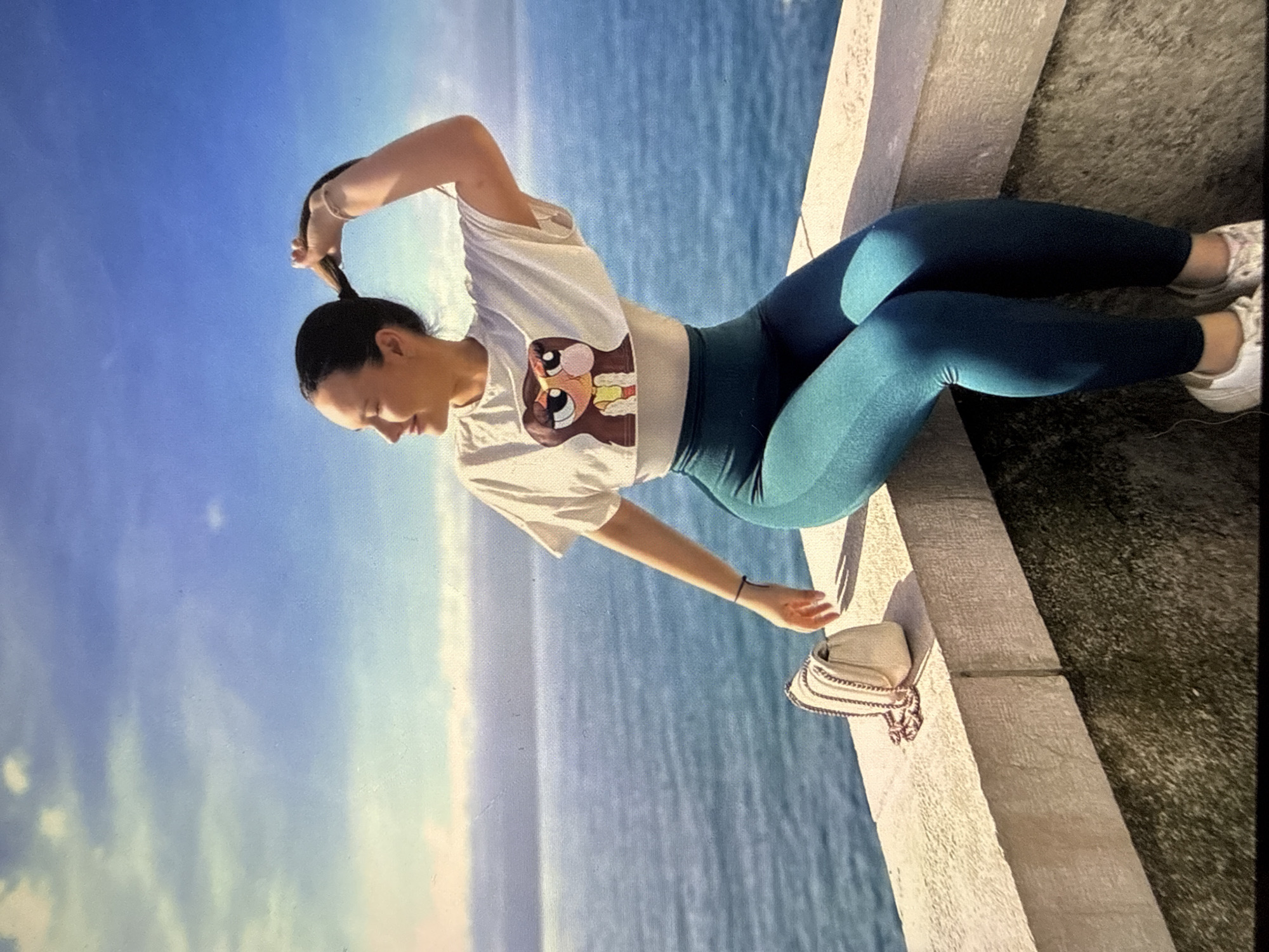 A woman is posing on a stone ledge. She is wearing a white t-shirt with a cartoon graphic, blue leggings, and white sneakers. She is pulling her hair into a ponytail. Next to her is a white purse. The background shows a body of water and a blue sky. While I cannot determine the exact location in Sofia, Bulgaria, based solely on this image, the presence of water and a stone structure suggests a location near a body of water within the city, such as a lake or reservoir along the outskirts of Sofia.