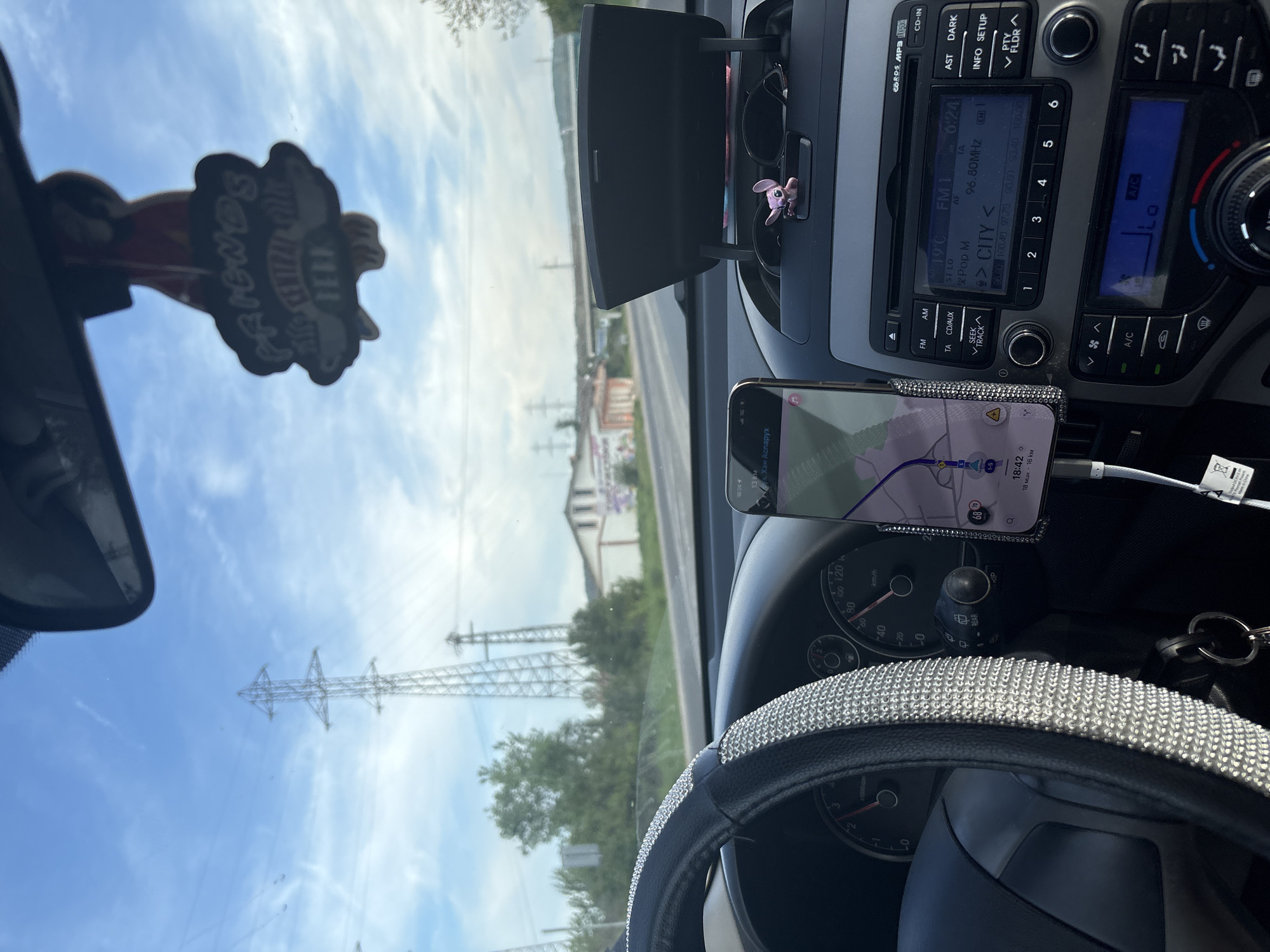 The image depicts the interior of a car, likely in motion. The dashboard is visible, including a car stereo tuned to FM1 96.80MHz, a GPS navigation system running on a smartphone, and a steering wheel with a decorative cover. Through the windshield, power lines and buildings are visible. The GPS indicates a location of approximately 13 km away from the destination "Хан Аспарух," in or near Tulovo, Bulgaria.