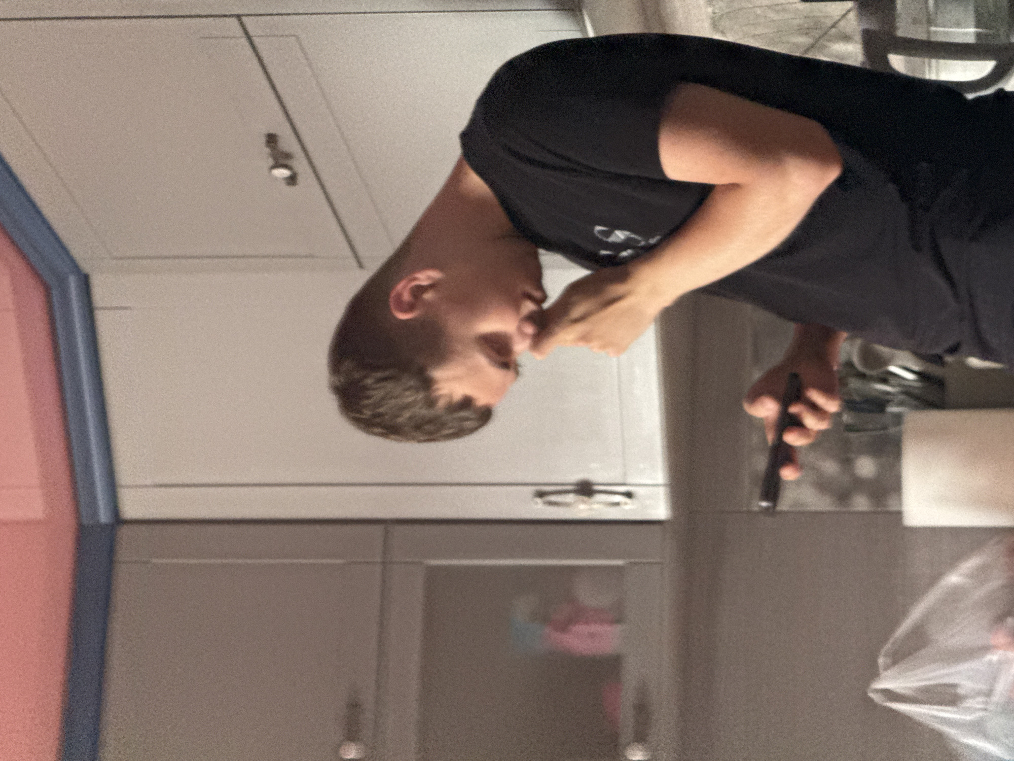 A young man is captured in an indoor shot, leaning forward with his head down, looking at his phone. He is wearing a black t-shirt. His right hand is raised towards his face, touching his nose. The background reveals a kitchen area with light-colored cabinets, one of which has a visible handle. The overall lighting suggests an indoor environment. The image does not provide any contextual information that would place it in Bohunove, Ukraine or any other specific location.