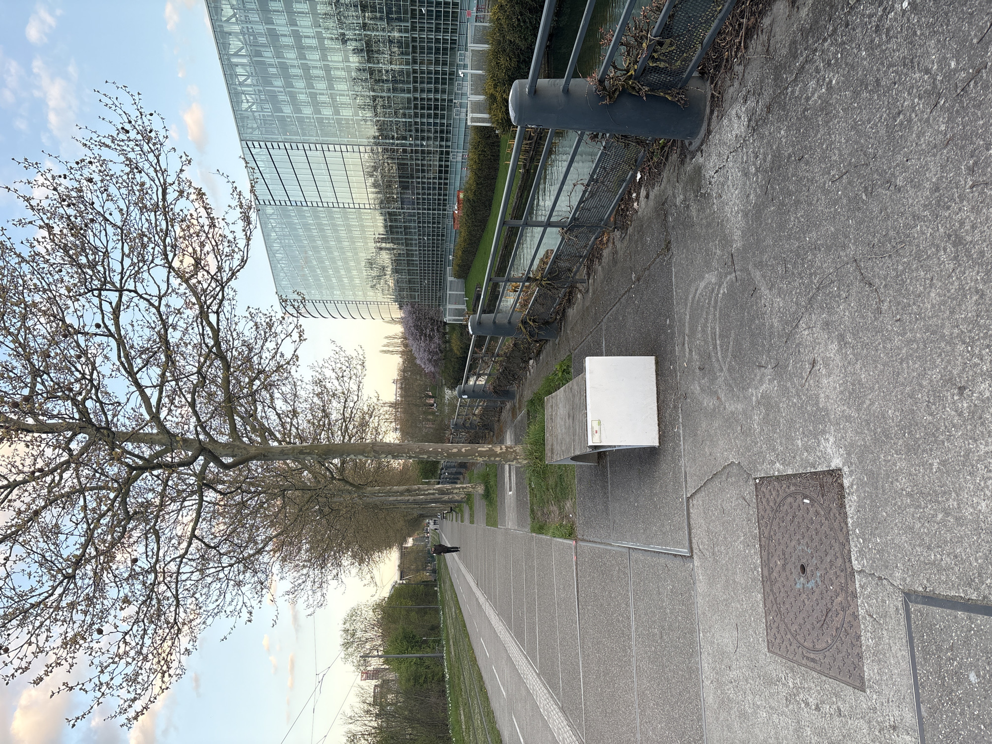 The image depicts a sidewalk along a canal in Strasbourg, France. In the background is a modern glass building, likely a government or office building. A metal railing separates the sidewalk from the canal. Trees line the sidewalk, some still bare from winter. A bench sits along the path. A person walks in the distance. The sidewalk is constructed from concrete slabs, with a manhole cover visible on the right.