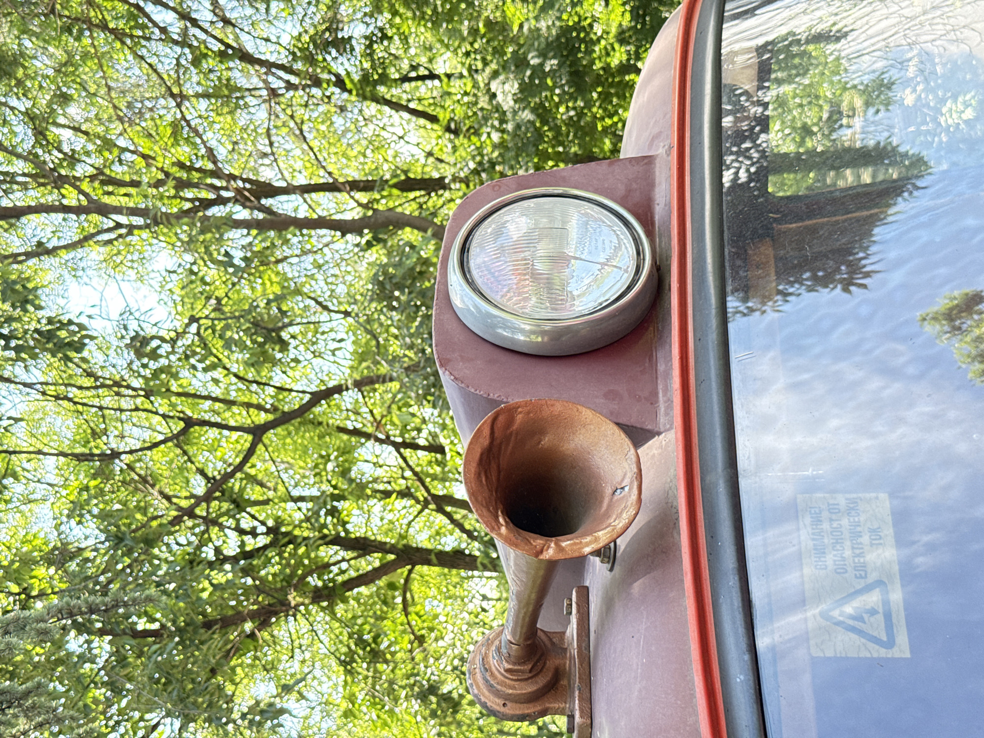 The image shows a close-up of the front of a vintage vehicle, likely a train or trolley, in an outdoor setting with trees in the background. The vehicle is maroon and features a round headlight with a silver rim and a bronze-colored horn below it. The vehicle's windshield is visible, with a warning sticker in Cyrillic script near the lower right corner. The location is likely Plovdiv, Bulgaria, given the Cyrillic script on the sticker. The trees in the background suggest a natural or park-like setting. There are no people visible in the image.