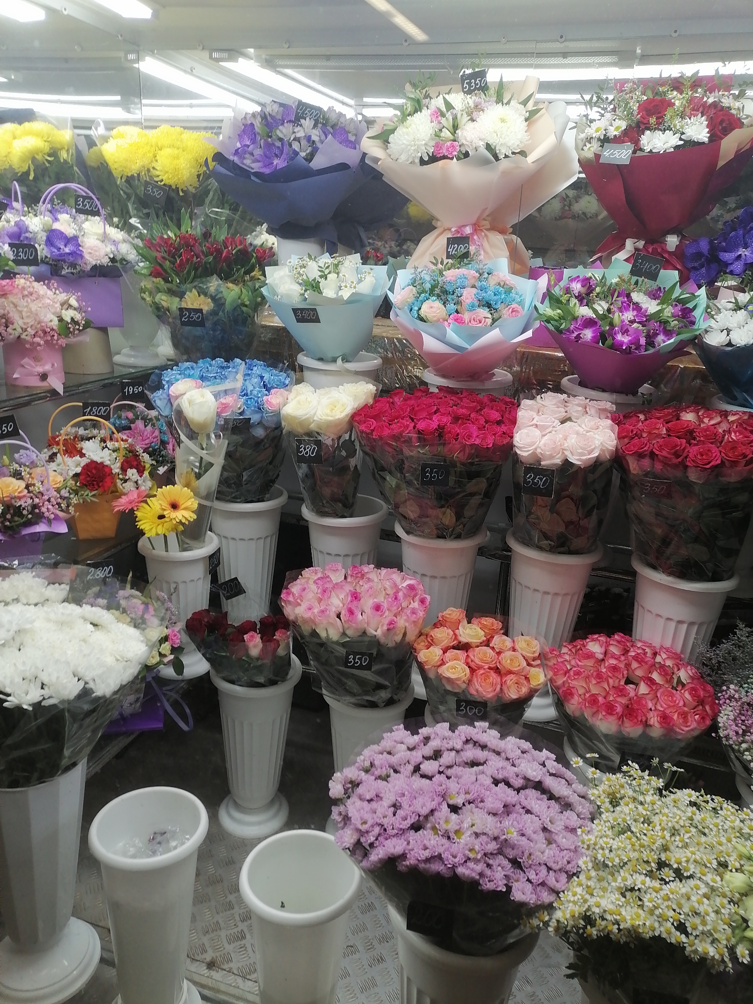 The image depicts a flower shop display in Moscow, Russia, featuring a wide assortment of bouquets and arrangements. The flowers are presented in various sizes and colors, including yellow chrysanthemums, purple irises, pink roses, white roses, red roses, and small white daisies. Many of the bouquets have price tags attached, with prices ranging from 350 to 5350. The display is well-lit, with overhead fluorescent lights visible. The overall impression is one of abundance and variety, typical of a professional flower shop.