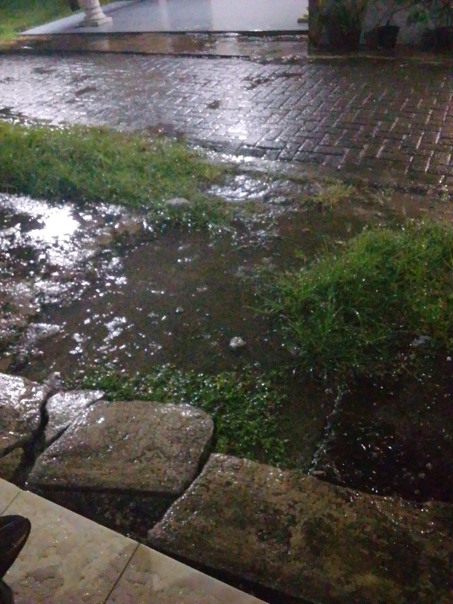 A wet paved pathway and grassy area are depicted, likely outdoors in Kedaidurian, Indonesia, suggesting recent rainfall. The ground is covered in puddles and reflections of light from an unseen source, indicating it is either dusk, nighttime, or overcast. The pathway appears to be made of interlocking bricks, and there are large, flat stones bordering a grassy patch. The grass is green and appears damp. A portion of a person's shoe is visible in the lower left corner of the image, suggesting their presence at the scene. The overall impression is one of a public outdoor space during or after a rain event.