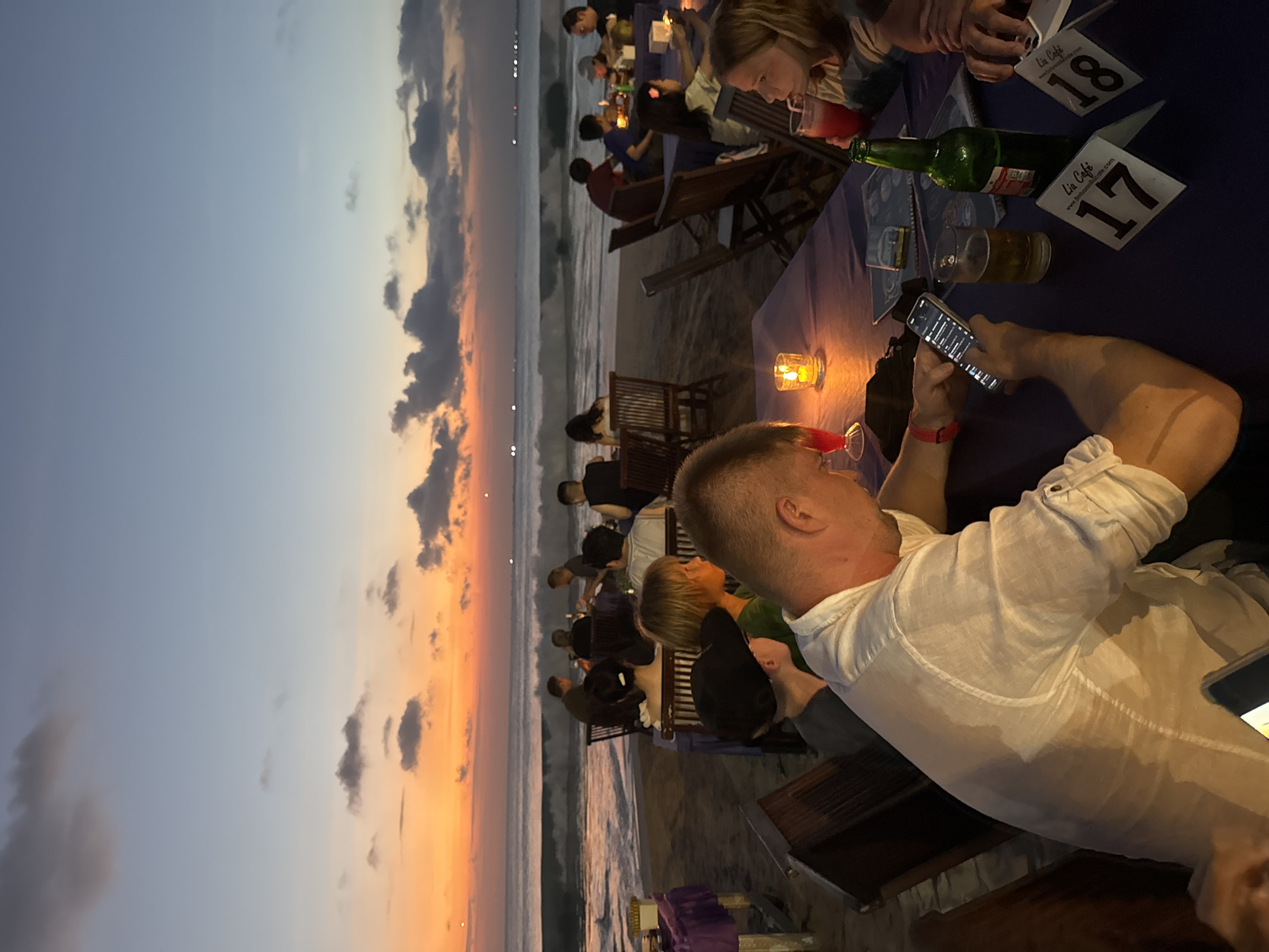 An outdoor dining scene unfolds on a beach in Kuta Selatan, Indonesia, during twilight. Several tables are set up on the sand, draped with purple tablecloths, each with candles providing ambient light. People are seated at these tables, some eating and drinking, while others are engaged with their phones. In the background, the ocean is visible with moderate waves and a sunset sky that transitions from orange to blue, dotted with scattered clouds. A few boats or distant lights can be seen on the horizon. The overall atmosphere is relaxed, suggesting a meal or gathering at a beachfront restaurant.