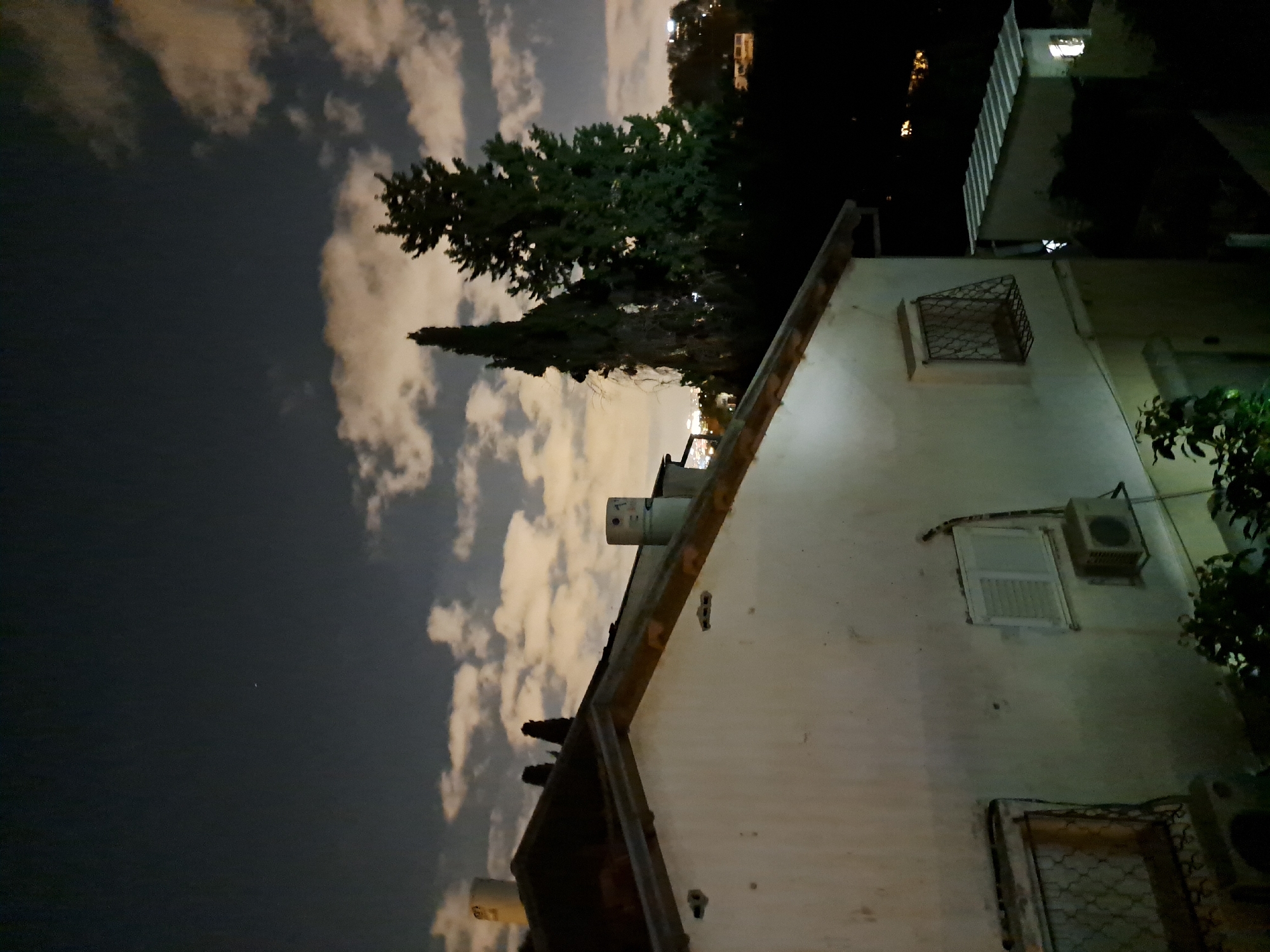 The image shows a night view of a residential area in Ramat HaSharon, Israel. In the foreground, a white, two-story house with a sloped, tiled roof is partially visible. The house has several windows, some with metal grates, and an air conditioning unit mounted on the exterior wall. Chimneys are present on the roof.
Behind the house, tall evergreen trees stand silhouetted against a dark sky dotted with scattered clouds illuminated by moonlight. In the middle ground, another smaller building with a corrugated metal roof is visible, with a single lit window. The background shows a blurred cityscape with distant lights, suggesting the location is within a suburban or urban environment. The overall impression is of a quiet night scene in a residential neighborhood.