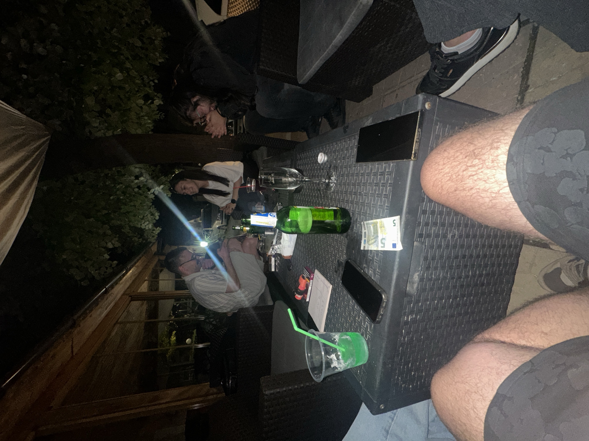 This image captures a night-time social gathering on an outdoor patio in Lom, Bulgaria. The scene is illuminated by artificial light, possibly from a phone flash, creating a stark contrast between bright and dark areas.
Several people are visible, seated around a dark, textured outdoor table. On the left side, two women are partially seen: one with long dark hair, wearing a white t-shirt, is smiling; another with dark hair and glasses, wearing a dark top, appears to be resting her head on her hands on the table, possibly laughing. A man wearing glasses and a striped shirt is seen on the lower left, illuminated by a bright light source, appearing engaged with something on the table. In the immediate foreground, the hairy legs of a person wearing dark shorts are visible, with one leg elevated towards the table. Another person's leg with a black sneaker and white sock is visible on the upper right side.
The table is cluttered with various items, suggesting a relaxed evening of drinks. Objects include an empty wine glass lying on its side, a green glass bottle (likely beer), and a clear plastic cup with a green drink residue and a green straw. Two black smartphones, a small white paper or notepad with faint, unreadable handwriting, and a cigarette pack or similar small box are also on the table. Notably, two 5 Euro banknotes are clearly visible.
The setting includes dark outdoor furniture, lush dark foliage in the background, and a light-colored awning or umbrella on the far left. The overall ambiance is casual and social, typical of an evening at an outdoor cafe or bar. The time of day is clearly night, evidenced by the darkness and reliance on artificial light.
