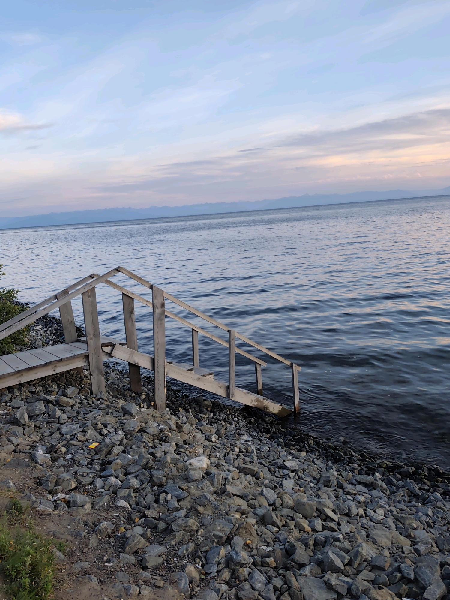 A wooden staircase leads from a rocky shore into a calm, dark body of water. The sky above is a gradient of pale blue and pink, with wispy clouds. Distant mountains form a hazy silhouette on the horizon. The scene appears to be a natural setting, possibly a lake or a large river, given the scale of the water and the mountainous backdrop. There is no vegetation visible around the immediate staircase area, only rocks. The overall impression is one of tranquility and natural beauty, possibly during sunrise or sunset.