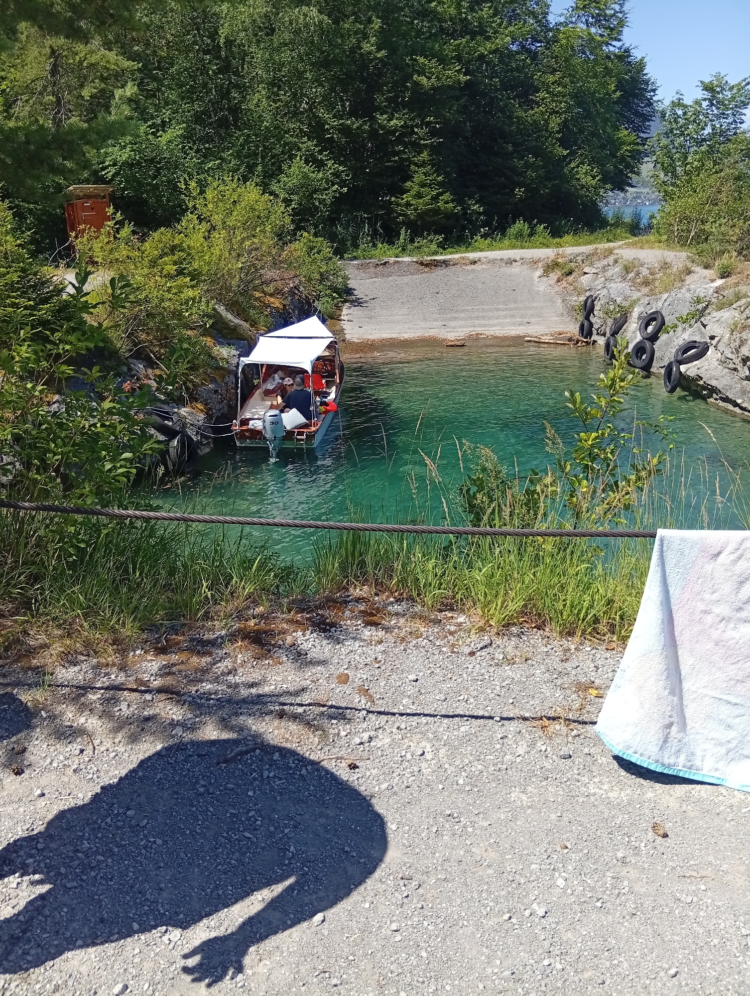 This is a daytime, eye-level, outdoor photograph taken at Emmetten, Switzerland, specifically at the boat ramp on Lake Lucerne, near the village's center. The image captures a scene of a small motorboat docked at the ramp, its white canopy casting a shadow on the clear, turquoise water. The boat appears to have several people on board. The ramp itself is made of concrete and gravel, leading into the lake. On the rocky bank to the right of the ramp, several old tires are stacked, presumably for protection. Lush green trees and foliage surround the area, and in the background, across the lake, a glimpse of distant buildings and mountains can be seen under a clear blue sky. In the foreground, a shadow of a person or object is cast on the gravel path, and a white towel is visible on the right side of the frame.