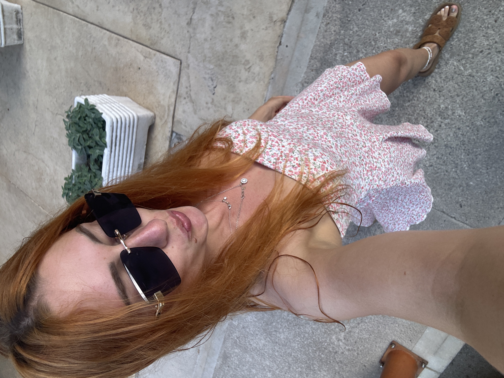 A woman with long, reddish-brown hair takes a selfie from a high angle, looking down into the camera. She wears large, dark, square-rimmed sunglasses with gold accents. Her lips are pursed. She is dressed in a light pink mini-dress with a small floral pattern and spaghetti straps. Multiple delicate silver necklaces adorn her neck. On her right ankle, she wears a white beaded anklet, paired with brown, open-toed sandals. Her right arm is visible, showing a small mole. The woman stands outdoors on a paved surface composed of light-colored, possibly concrete or stone, tiles with visible grout lines. In the background, two white, ribbed planters containing green leafy plants are visible. The scene is located in Budva, Montenegro.