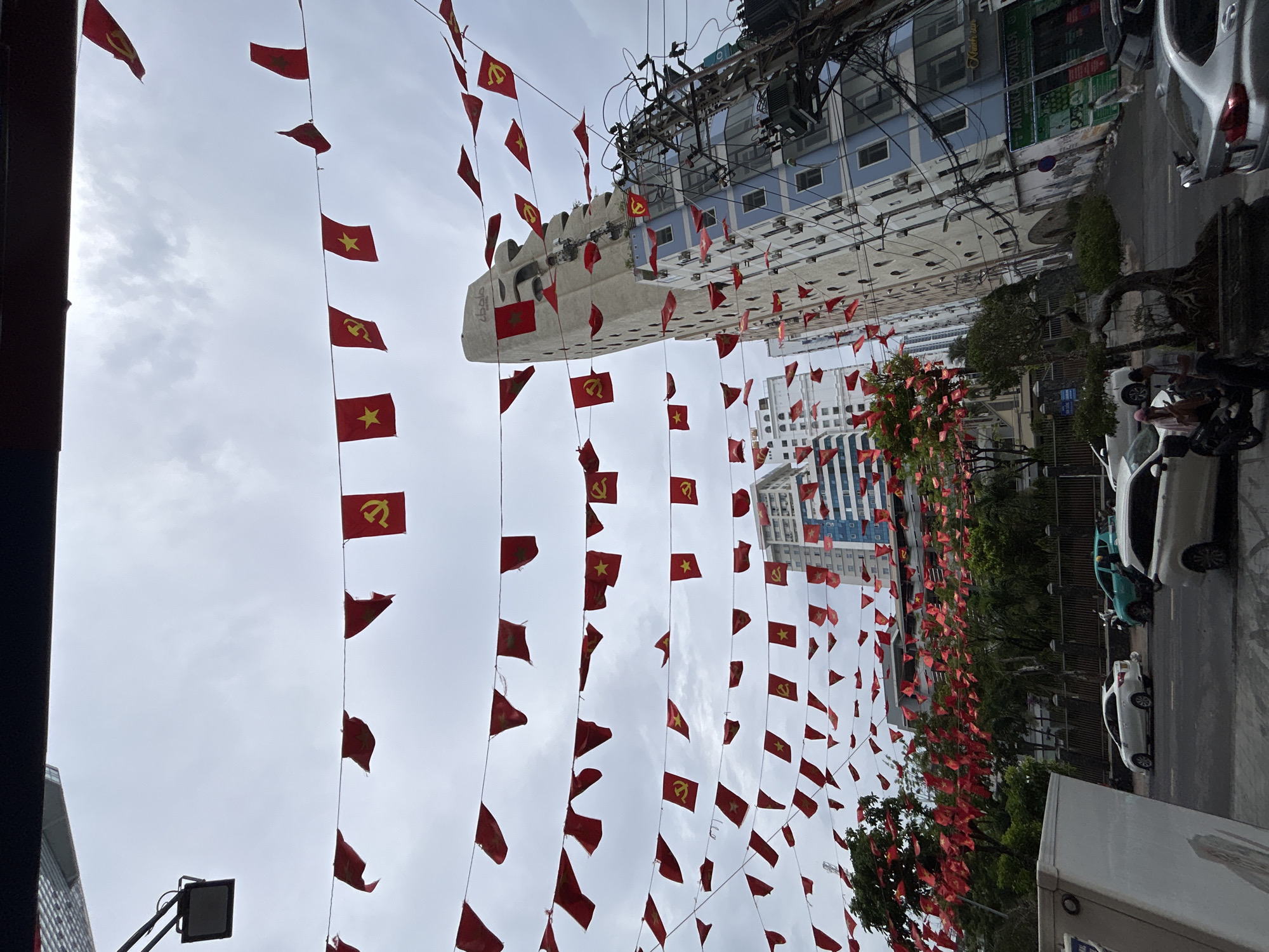 The image shows a street scene in Nha Trang Ward, Vietnam. Numerous small red flags with yellow stars or hammer-and-sickle symbols are strung across the street. Buildings line the street, including a blue and white building and taller buildings in the background. Trees are visible along the side of the street. Vehicles, including cars and scooters, are parked on the side of the road. People are visible near the scooters. The sky is overcast.