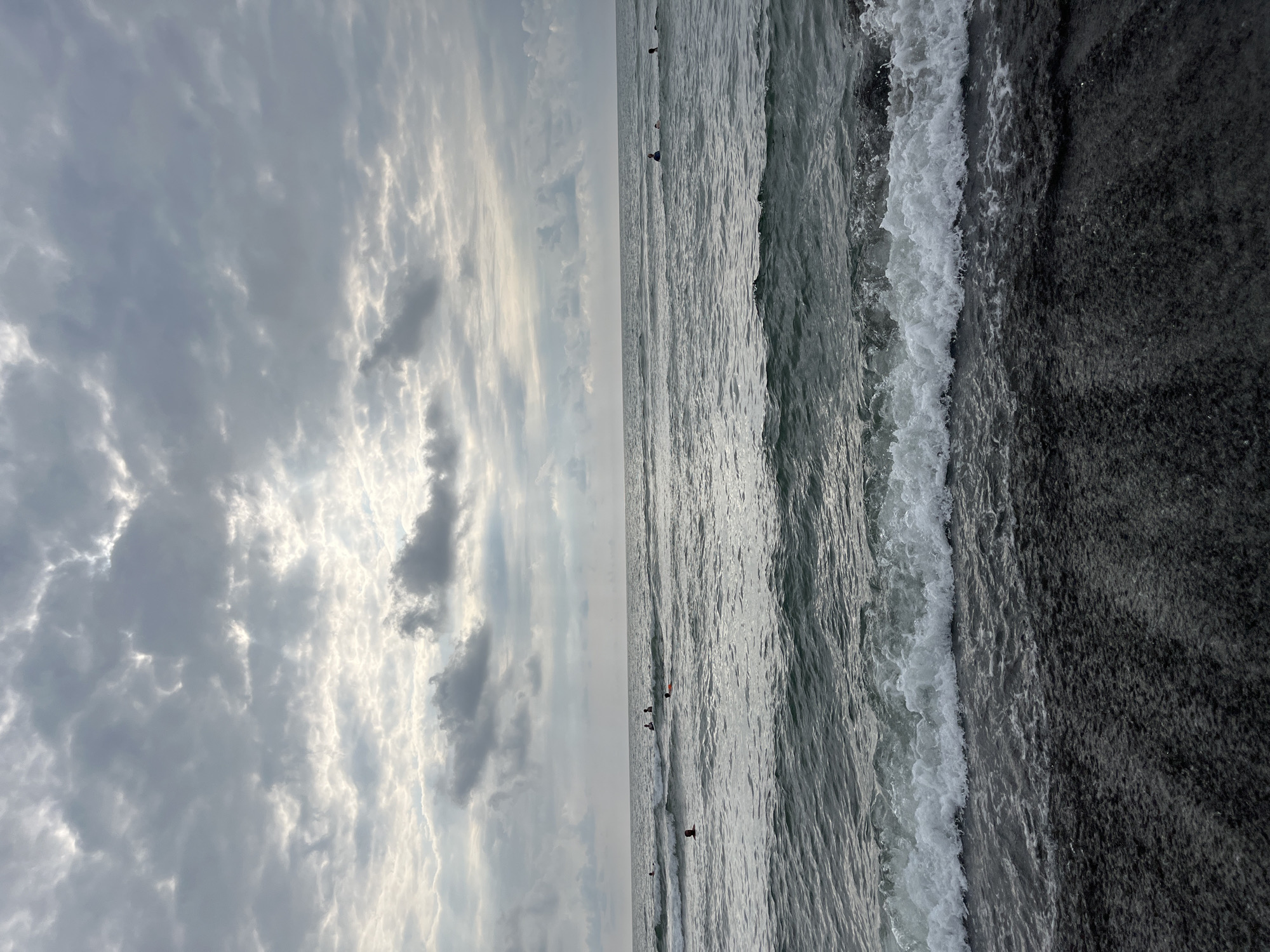 The image depicts a cloudy day at the Black Sea coast in Ureki, Georgia. The foreground shows dark, wet pebbles and sand, with a gentle wave breaking on the shore. The middle ground consists of the sea, with several people visible in the water. Some appear to be swimming, while others are likely surfers waiting for waves, with their boards partially submerged. In the background, the horizon is a clear line between the sea and the sky. The sky is filled with a dense layer of grey and white clouds, with patches of brighter light suggesting the sun is diffused behind them. The overall scene conveys a sense of a calm, overcast day at the beach.