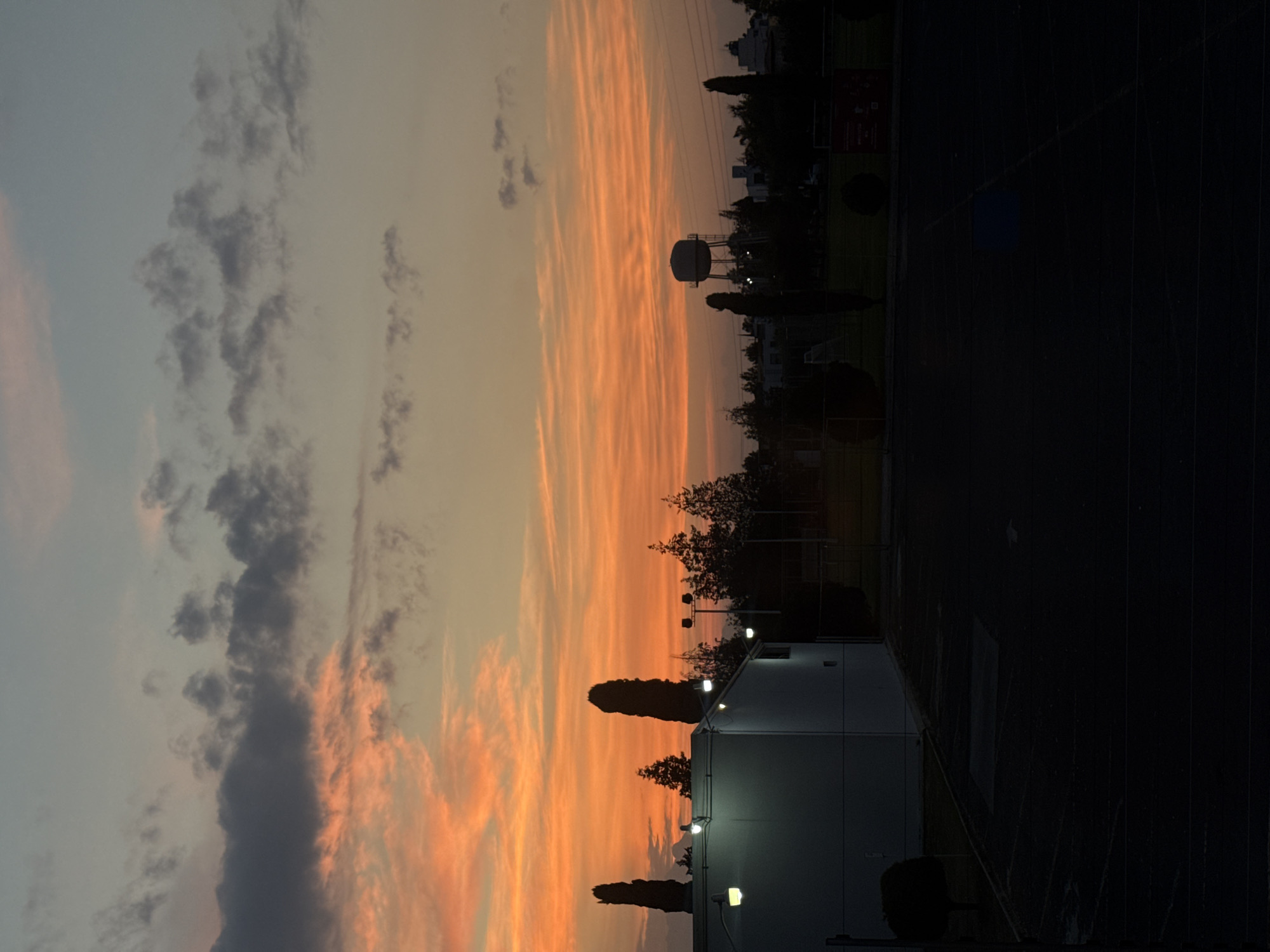 The image captures a twilight scene in Heroica Puebla de Zaragoza, Mexico. In the foreground, a white building is illuminated by several lights. Adjacent to the building is a dark running track and athletic field. In the distance, a water tower is visible, positioned amongst trees. The sky above the treeline is filled with orange-colored cirrus clouds. A smaller band of gray clouds is on the opposite side of the sky, toward the bottom left.