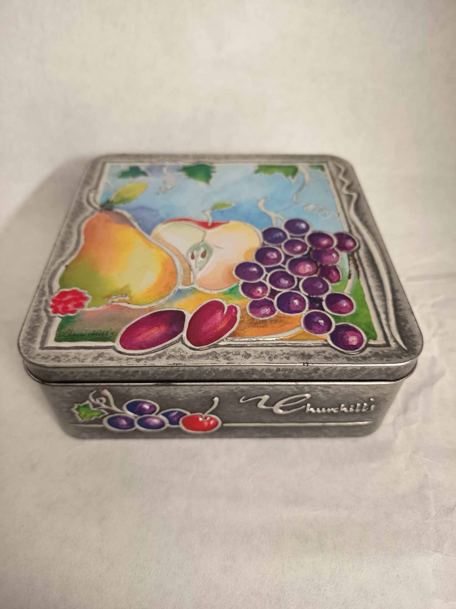A close-up shot depicts a metal tin with a rectangular shape and a slightly tarnished, silver-grey finish. The lid of the tin is adorned with a colorful illustration of various fruits including a pear, apple, plums, and a bunch of grapes, set against a background of blue and green hues. The design is framed by an embossed, decorative border. The side of the tin is partially visible, revealing the word "Churchill's" in a cursive font, with a decorative sprig of grapes with a cherry motif preceding the text. The tin is resting on a surface of off-white, slightly textured material. There are no visible people, actions, or interactions. The environment is sparse and without distinct location details or indicators of a city. The location of Wangen bei Olten, Switzerland is not identifiable from the image itself.