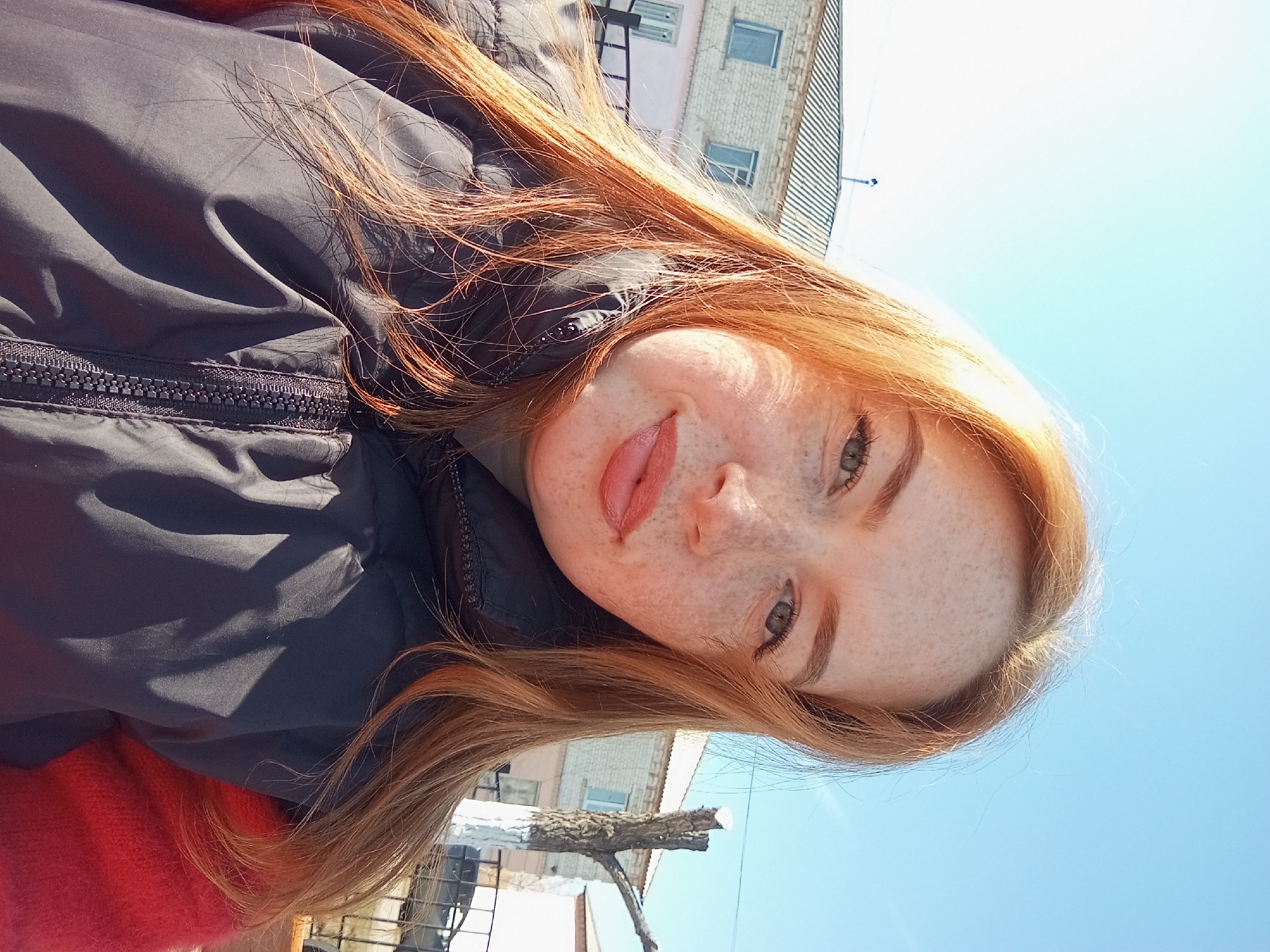 A close-up, tilted, outdoor selfie shows a young, light-skinned woman with freckles, green eyes, and reddish-orange hair sticking her tongue out playfully. She wears a black jacket with a visible zipper. The background includes a clear blue sky, part of a brick building with windows, a tree branch, and metal fencing. Based on the location information, this image was taken in Yurgamysh, Russia.