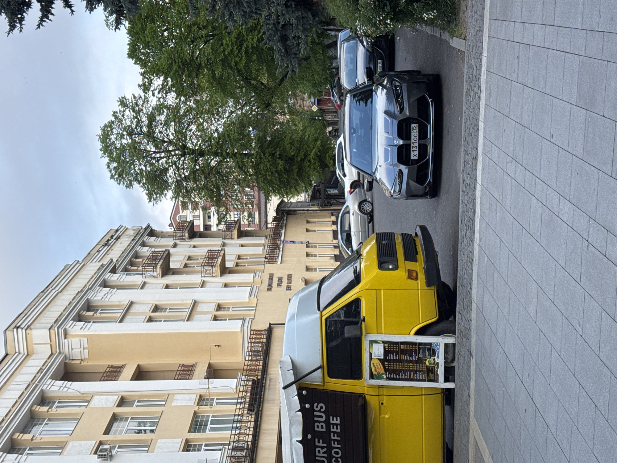 The image shows a street scene in what appears to be Akhieli, Georgia. A yellow van with "SURF BUS COFFEE" signage is parked along the right side of the street, adjacent to a gray tiled sidewalk. Its menu is visible. Parallel to the van, on the road, are several parked cars, including a gray BMW with the license plate "X 131 OC 15". A light-colored, multi-story building with multiple windows and dark iron balconies is on the left. Overhanging trees are present in the center and top sections of the image. The sky is overcast.