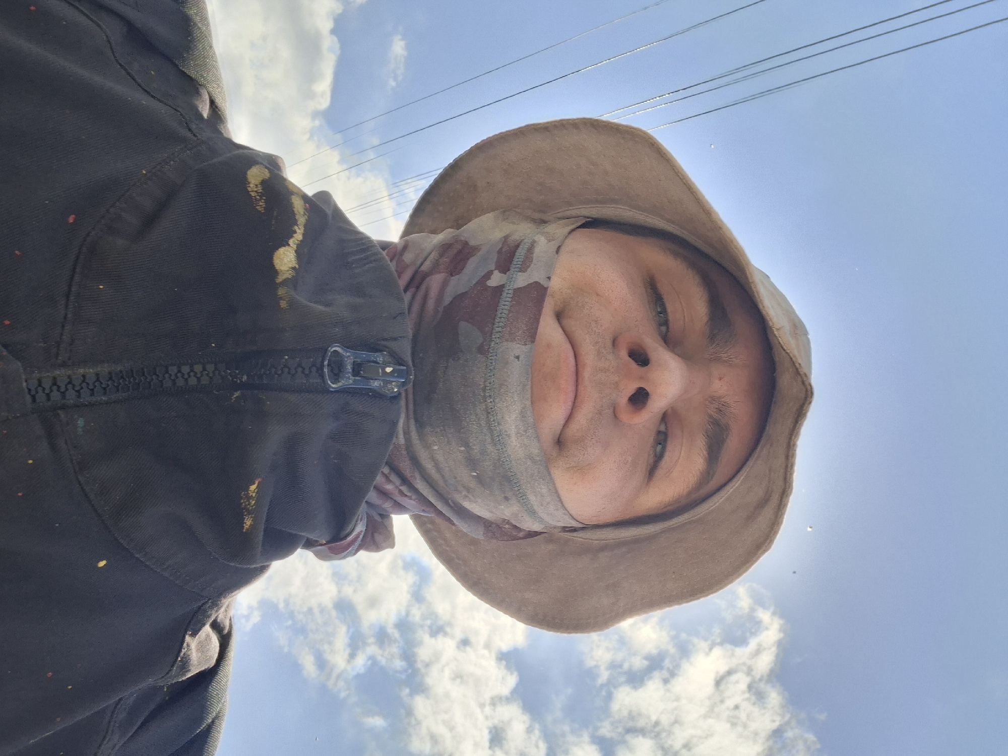 A man in a beige wide-brimmed hat and camouflage neck gaiter takes a selfie outdoors, angled upwards towards the sky. He's wearing a dark-colored jacket or work suit with a zipper and some paint splatters. The background consists of a blue sky with scattered white clouds and thin electrical wires overhead. The location is Unknown, Unknown.