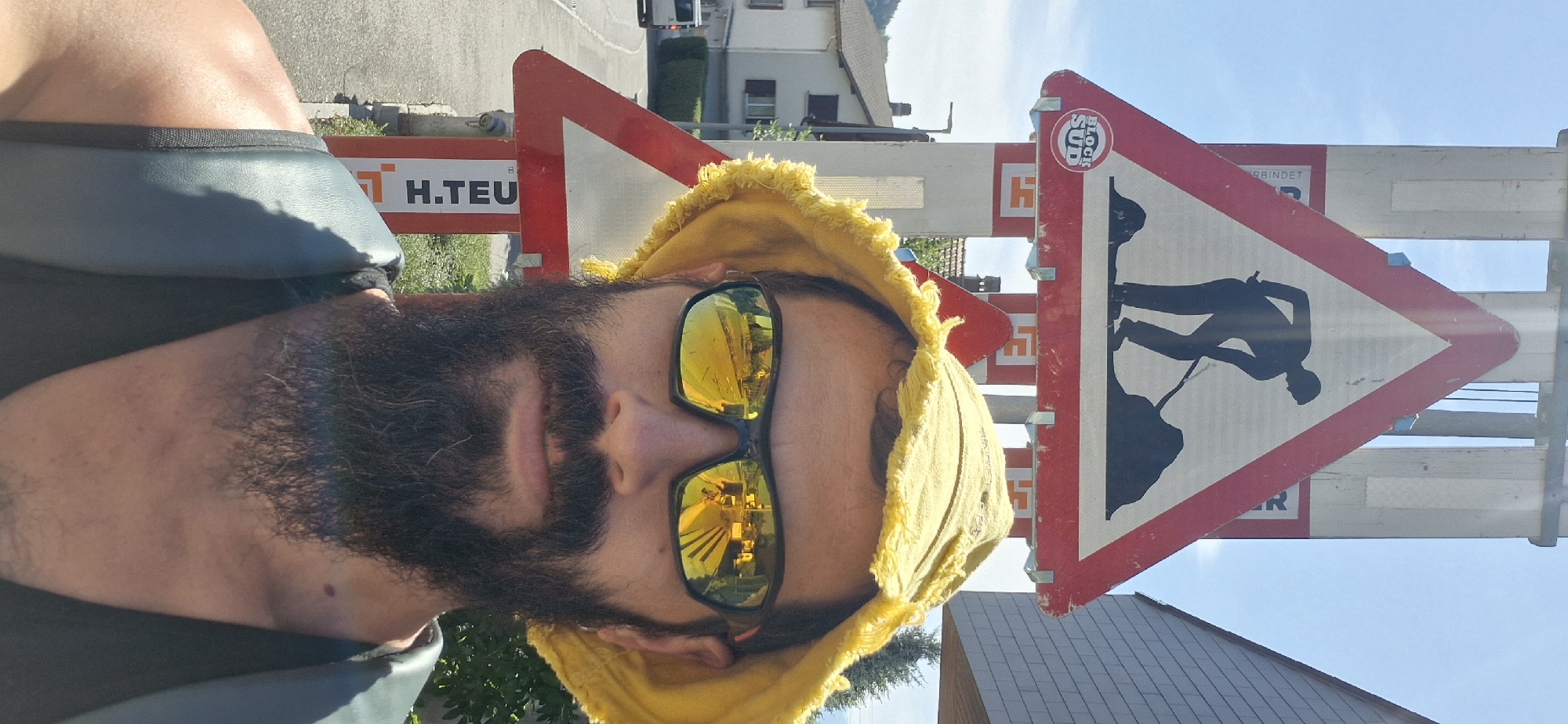 This media file features a close-up, tilted selfie of a person with a full beard and sunglasses. They are wearing a yellow bucket hat with frayed edges. The background shows a road with construction signs and some buildings. The sun is shining brightly, suggesting it is daytime, and the weather appears to be good.
The construction signs indicate road work is in progress. One sign is a triangular warning sign with a red border and a white interior, depicting a silhouette of a person digging. Another sign is rectangular and reads "H.TEU" in black text on a white background with a red border. A smaller circular logo with the text "BLOCK SUD" is also visible on one of the signs. The buildings in the background suggest a semi-urban or rural setting, possibly in Wattenwil, Switzerland, as suggested by the context. The reflection in the sunglasses shows a bright, sunny environment, possibly a street or highway with vehicles. The overall mood seems casual and relaxed, despite the presence of road construction.