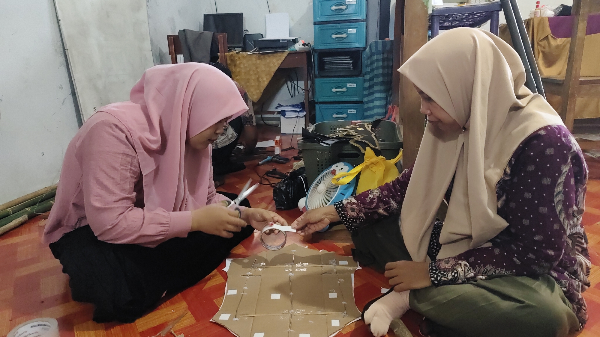 In a dimly lit room with wooden floors, two individuals are seated on the floor engaged in a crafting activity. The person on the left, wearing a pink hijab and a pink shirt, is using scissors to cut a strip of white tape. Their hands are close to a piece of cardboard that is laid out on the floor, marked with white squares and lines. The person on the right, wearing a beige hijab and a purple patterned shirt, is also holding a strip of white tape, assisting in the taping of the cardboard. The cardboard appears to be a component of a larger project, possibly a costume or prop, given its shield-like shape. The background reveals a cluttered environment with various items, including a desk with a computer monitor and printer, blue plastic drawers, a laundry basket filled with black fabric, a small fan with a cartoon character on it, and other assorted materials, suggesting a workshop or personal crafting space. The scene is set within a residential building in Banyubunih, Indonesia, a small village known for its traditional crafts.