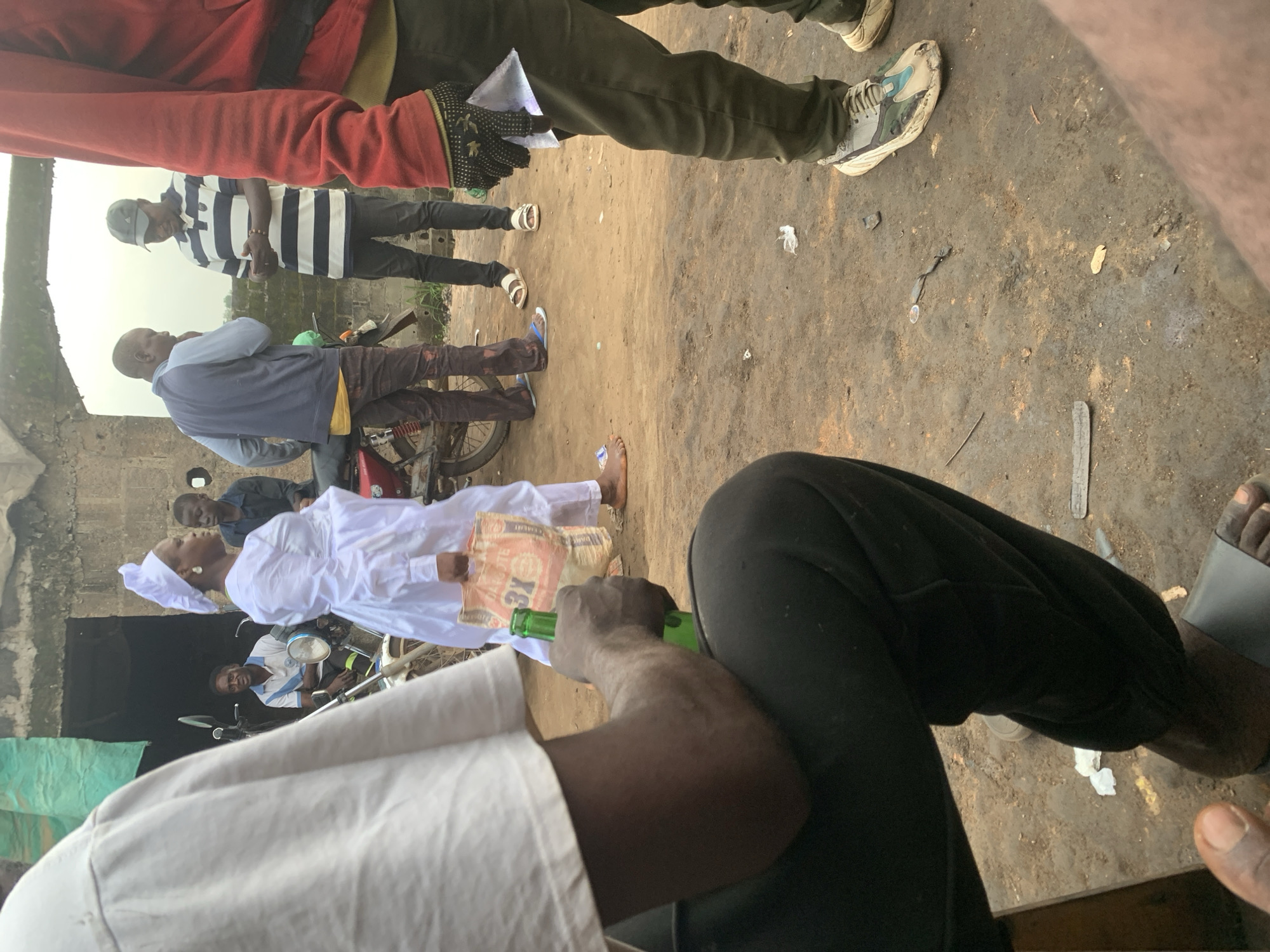 In Shagamu, Nigeria, several individuals are gathered outdoors. In the foreground, a person's arm is visible holding a green glass bottle. Behind them, a woman in a white dress and head covering carries a large orange bag. To her right, a young boy in a dark shirt stands near a motorcycle. Further back, two men converse; one wears a blue and gray jacket and the other, a striped shirt and a baseball cap. Another person is partially visible on the far right, wearing a red jacket and dark pants. The ground is dirt and appears somewhat dry. A motorcycle is parked in the middle ground. The environment suggests an informal gathering or street scene.