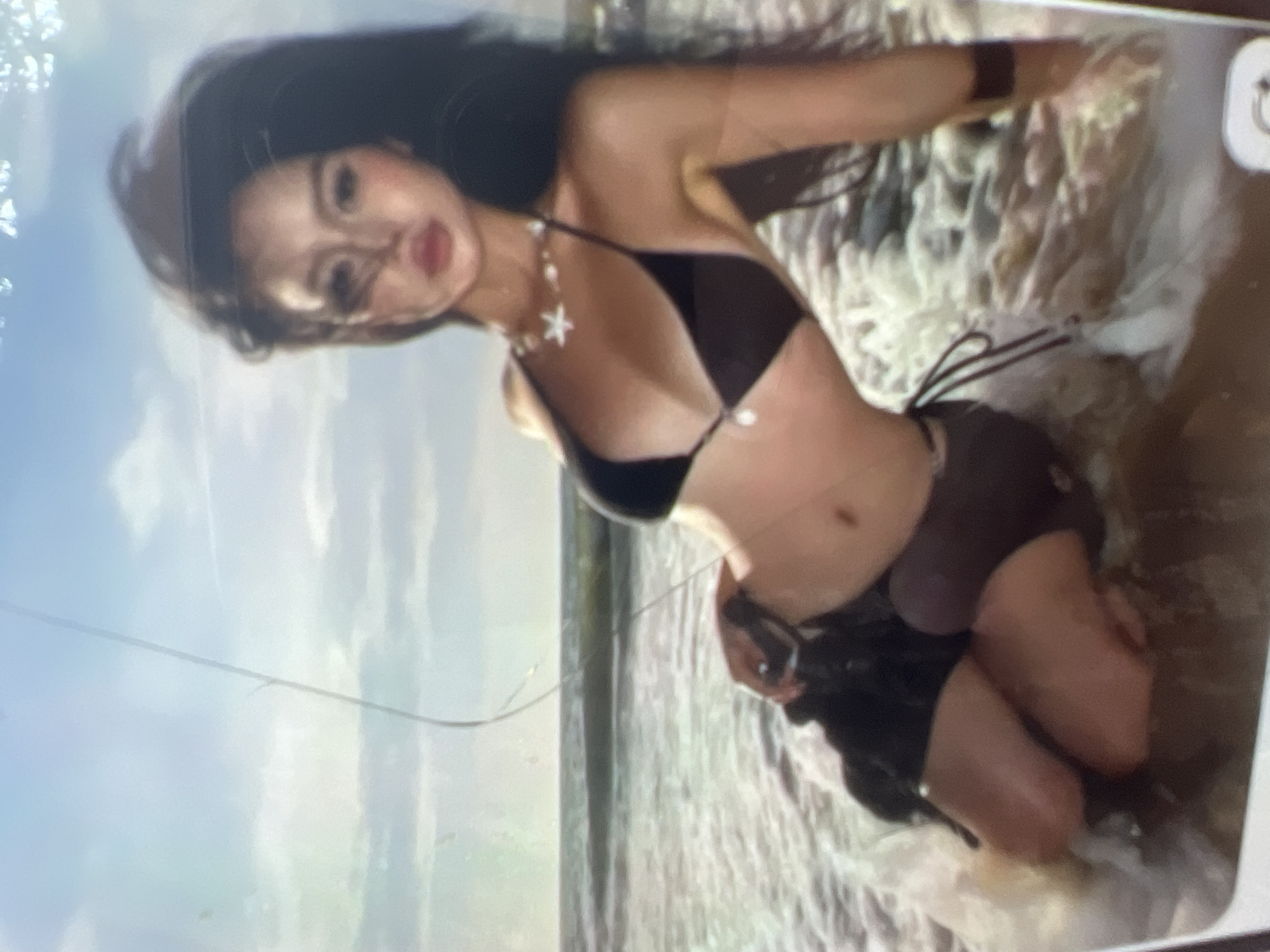 A young woman is kneeling in shallow water near a beach. The woman has long dark hair and is wearing a dark bikini and dark shorts. She is looking towards the viewer with her head slightly tilted. Her body is angled to the left of the frame. The water is foamy and surrounds her legs. The background consists of a body of water with waves and a sky with clouds. There is a thin, vertical line across the image, possibly a scratch on a screen or a thin object in the foreground. The image appears to be a photograph. The location of Kara-Dıykan, Kyrgyzstan is not discernible from the image.