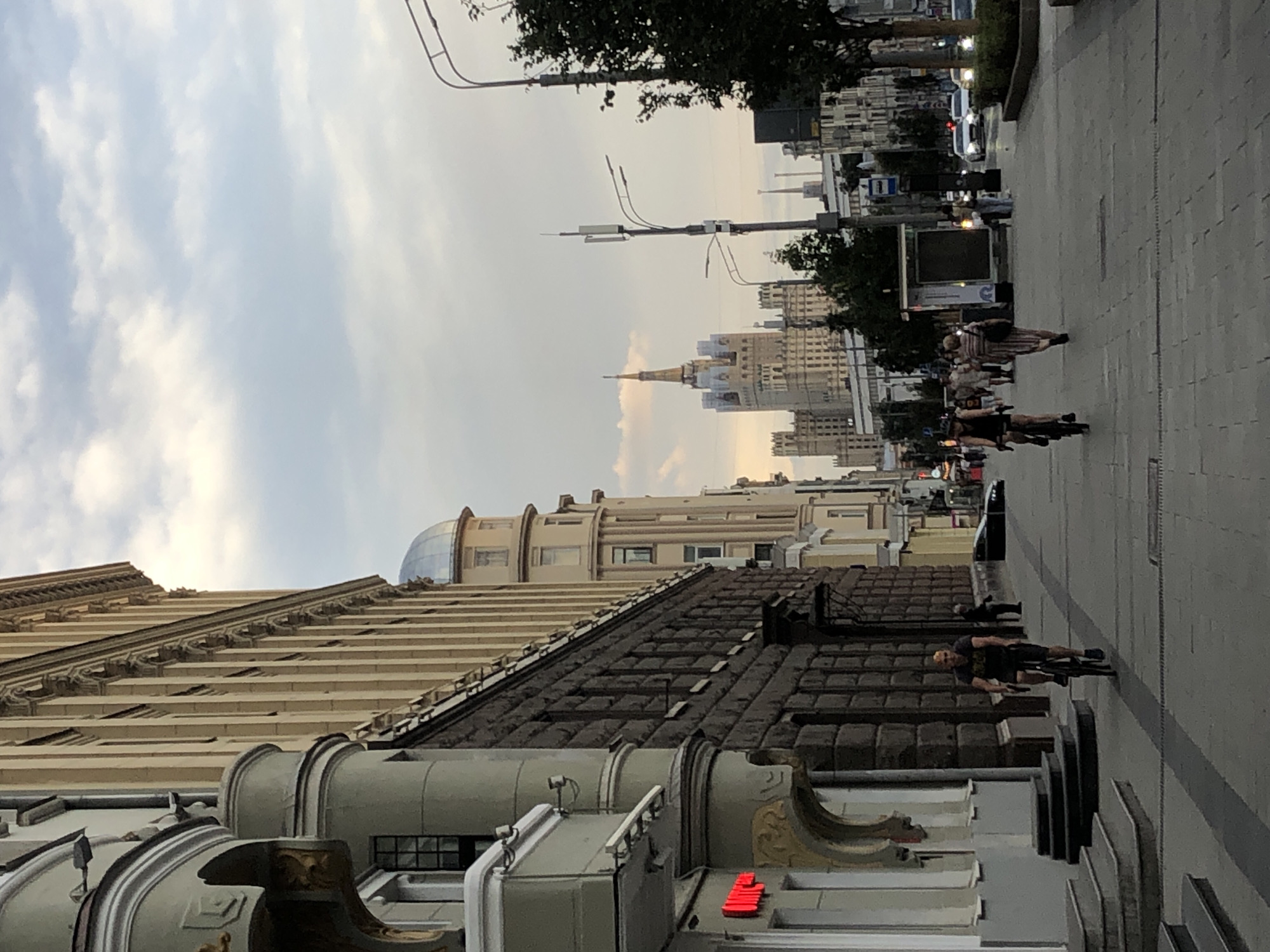 The image captures a street scene in Moscow, Russia, under a cloudy sky. On the left, a grand building with ornate architectural details, including a curved facade and decorative elements, dominates the foreground. The building features a dark stone lower level and lighter colored upper floors with vertical lines. Further down the street, other buildings with similar architectural styles are visible, including one with a domed roof and another with a distinctive spire. A prominent Stalinist skyscraper, known as one of the Seven Sisters, stands in the distance.
Several people are present on the paved sidewalk. Two individuals are riding bicycles, one in the mid-ground and another further ahead. Other pedestrians are walking, with one woman in a striped dress walking away from the camera and a few others in the distance. Cars are visible on the street, suggesting it is a moderately busy urban area. Streetlights and utility poles are also present, lining the pedestrian walkway and the street. The overall impression is of a European city street during daylight hours, possibly in the late afternoon or early evening, with a mix of historical and modern architecture.