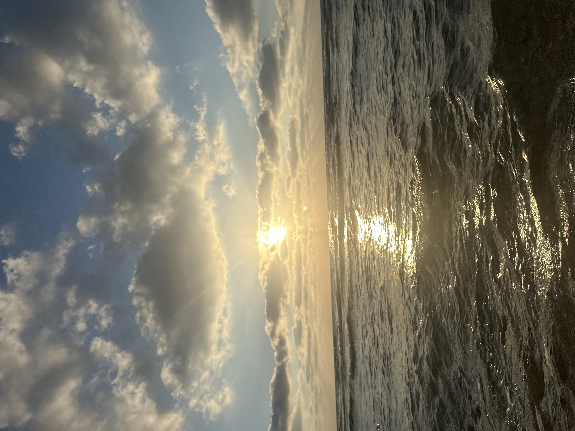 This is a vertical, eye-level, outdoor photograph of the sea and sky at sunset. The sun is low in the sky, casting a warm glow and rays of light through scattered clouds. The water is dark and choppy with reflections of the sun on the surface. The scene is likely a beach in Haifa, Israel, given the characteristic Mediterranean light and sea conditions. There are no people or objects visible in the image, focusing solely on the natural landscape.