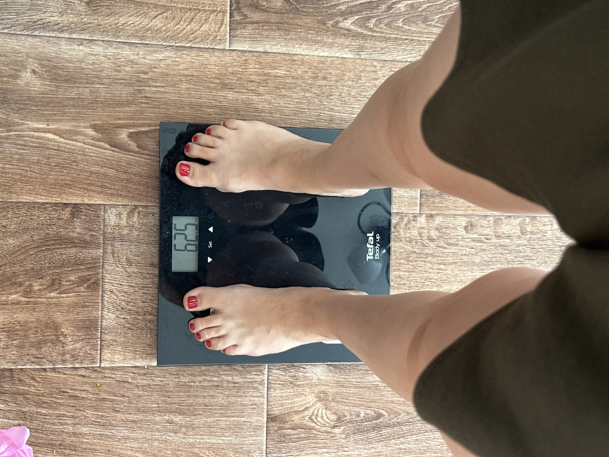 A person's feet and lower legs are shown standing on a black digital scale, which is placed on a wooden floor. The scale displays the number "62.5". The person has red nail polish on their toes. The background consists of a light brown wooden floor with visible grain patterns. The image is taken from a top-down perspective. The location is not specified in the image or the provided crops.