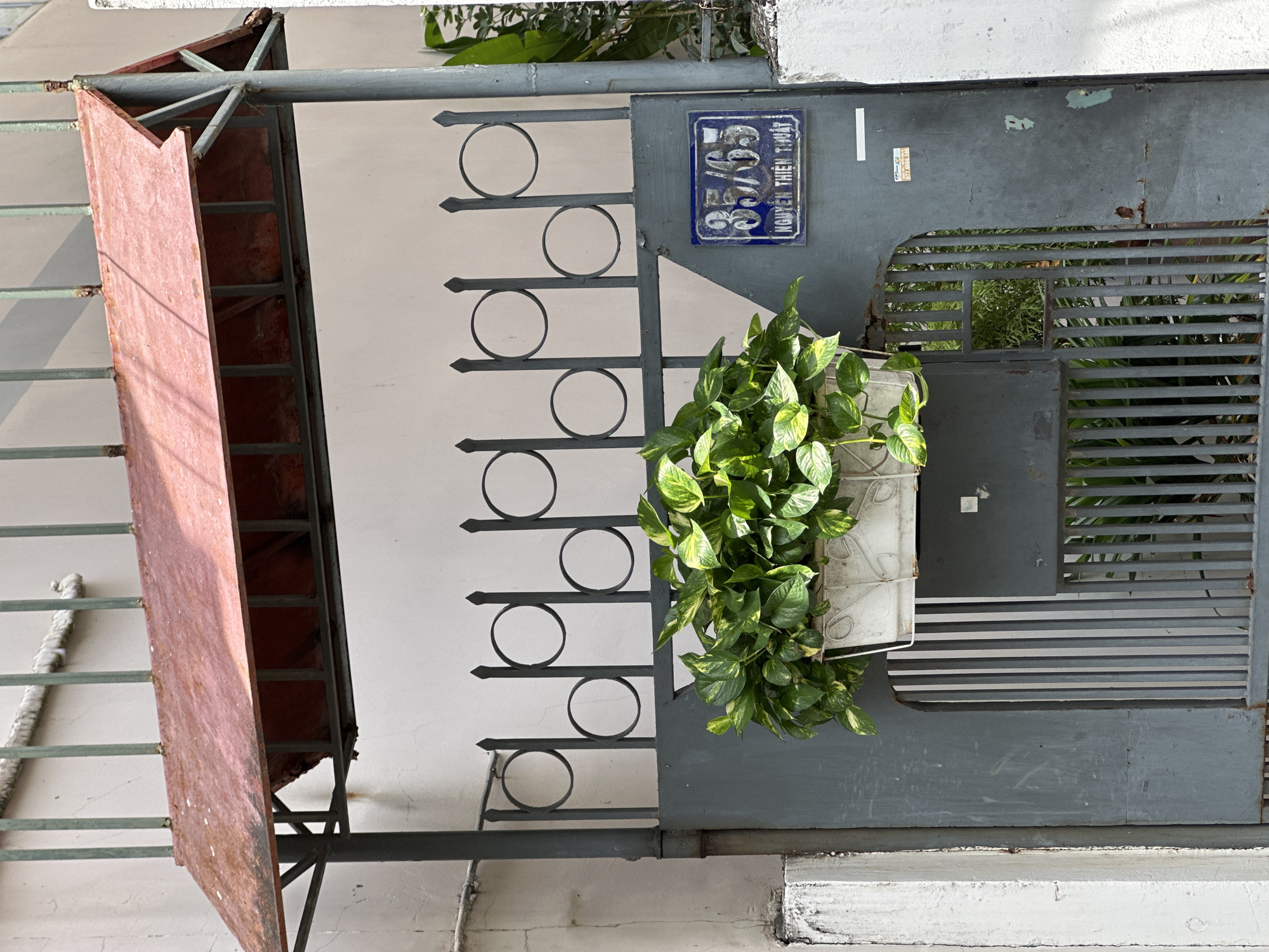 The image shows a close-up of a residential gate in Nha Trang Ward, Vietnam. The gate is made of dark gray metal and features a decorative pattern of circles and vertical bars. A planter box filled with lush green plants hangs from the top of the gate. To the right of the plants, a blue ceramic house number plaque displays "35/65" in white numerals, with the street name "NGUYEN THIEN THUAT" written below. A red metal awning with visible supports is situated above the gate. The surrounding wall is light-colored and appears weathered. The overall scene depicts an entrance to a home, suggesting a typical urban residential setting.