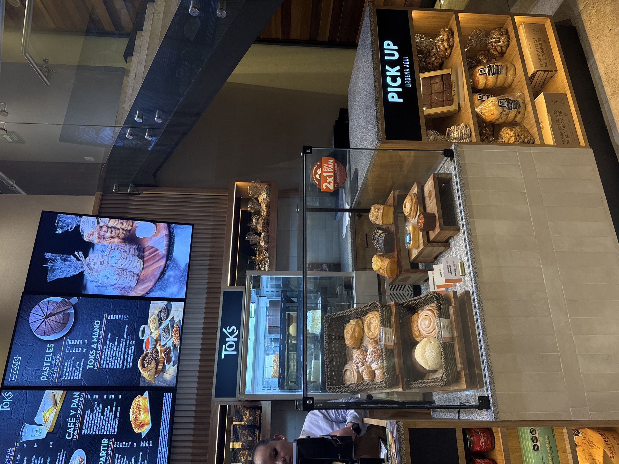 The image depicts the interior of a Toks restaurant in Mexico City, Mexico. A menu board on the left lists various food and drink options, including "pasteles" (cakes), "café y pan" (coffee and bread), and "productos artesanales" (artisanal products). Display cases showcase pastries and baked goods, including croissants, cinnamon rolls, and loaves of bread. A "Pick Up" sign indicates an area for order collection. There are employees visible, presumably preparing orders or assisting customers.