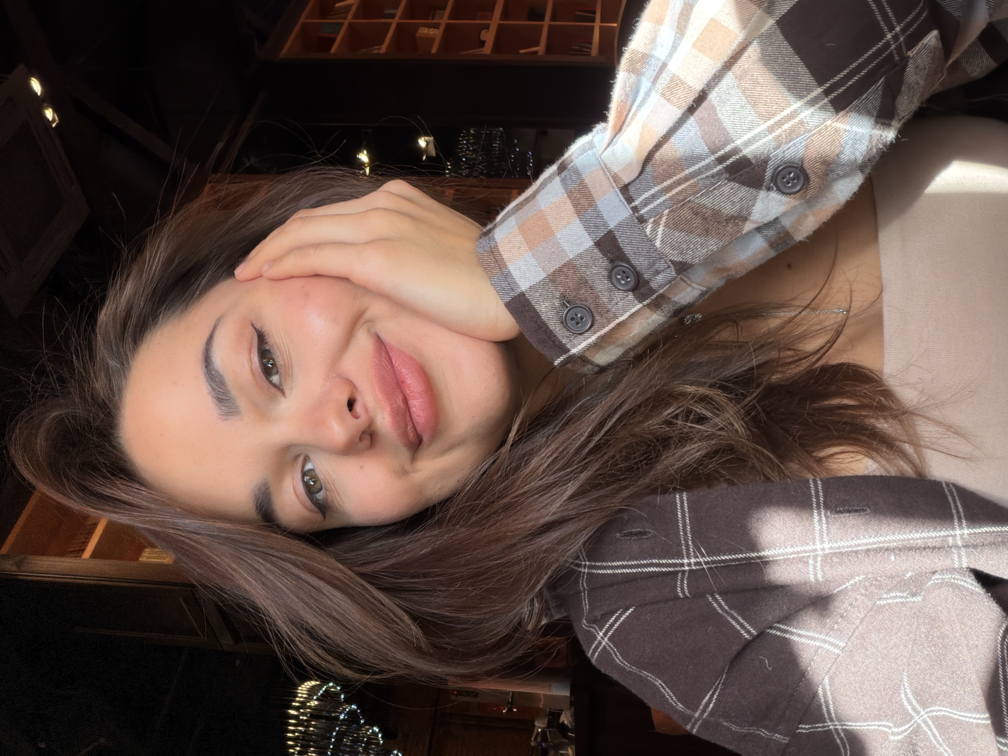 A young woman poses indoors. She has long brown hair, light skin, and is wearing a plaid shirt. Her left hand is touching her face. The background includes a shelf with objects and blurred lights. The location of the photo is not discernible from the image. The photo cannot be verified as being taken in Krasnoyarsk, Russia.