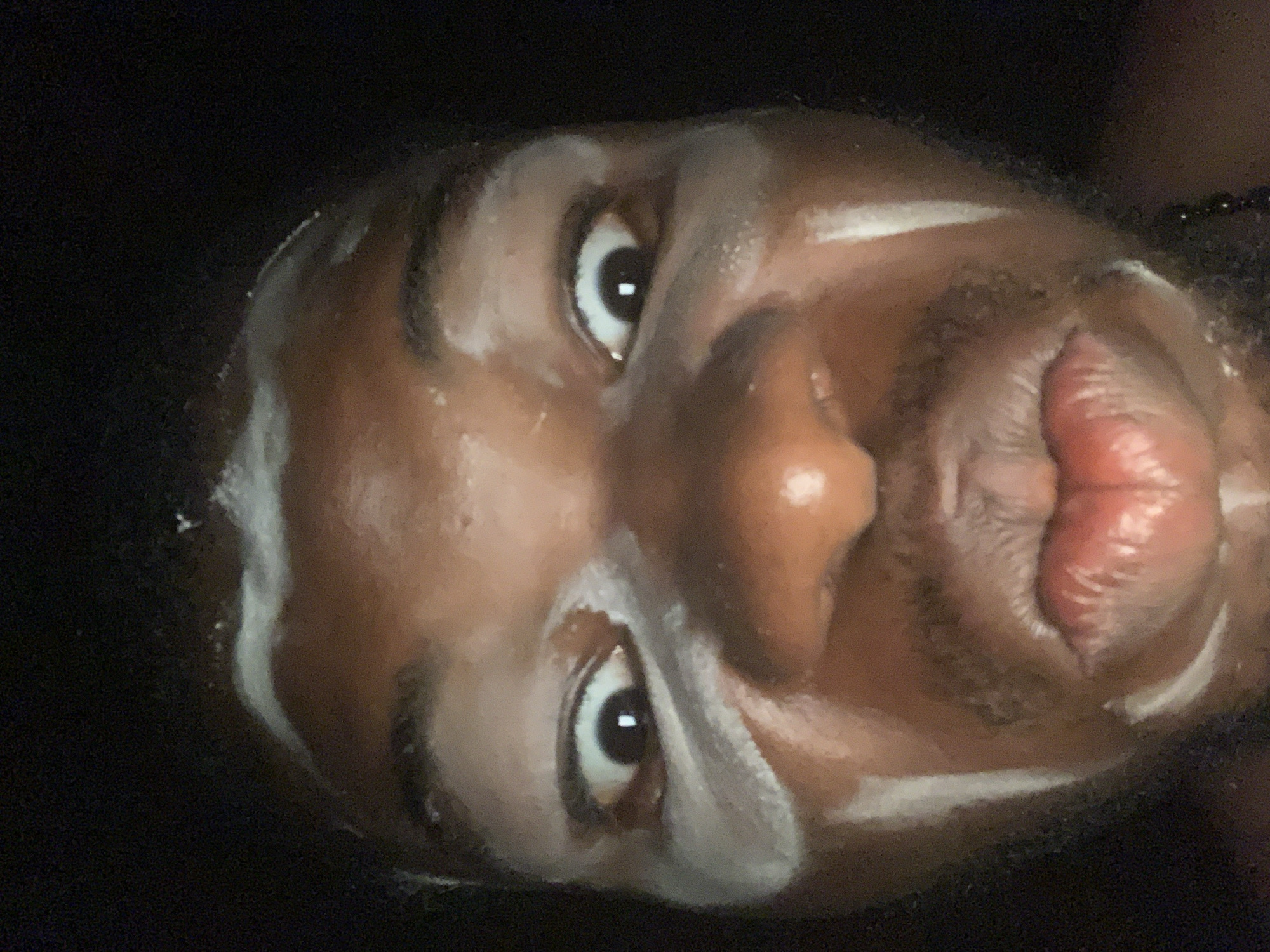 A close-up, upside-down image shows a person with dark skin and facial hair. White cosmetic product is applied around the eyes and on the forehead and chin. The person's eyes are open, and their lips are pursed. The background is dark. No information can be inferred about the location of Onitsha, Nigeria.