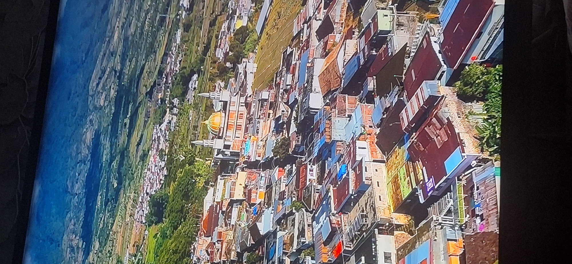 This is an aerial view of a densely populated town or city, likely located in a hilly or mountainous region, with a prominent building featuring a golden dome and two minarets, resembling a mosque or a religious structure. The architecture of the surrounding buildings suggests a warm climate, with many houses having flat or slightly sloped roofs, some made of corrugated metal. The terrain is terraced, with agricultural fields visible in the background and on the slopes. There are no visible people or discernible actions. The sky is clear and blue, suggesting a sunny day. There is no indication that this location is in Bila Tserkva, Ukraine.