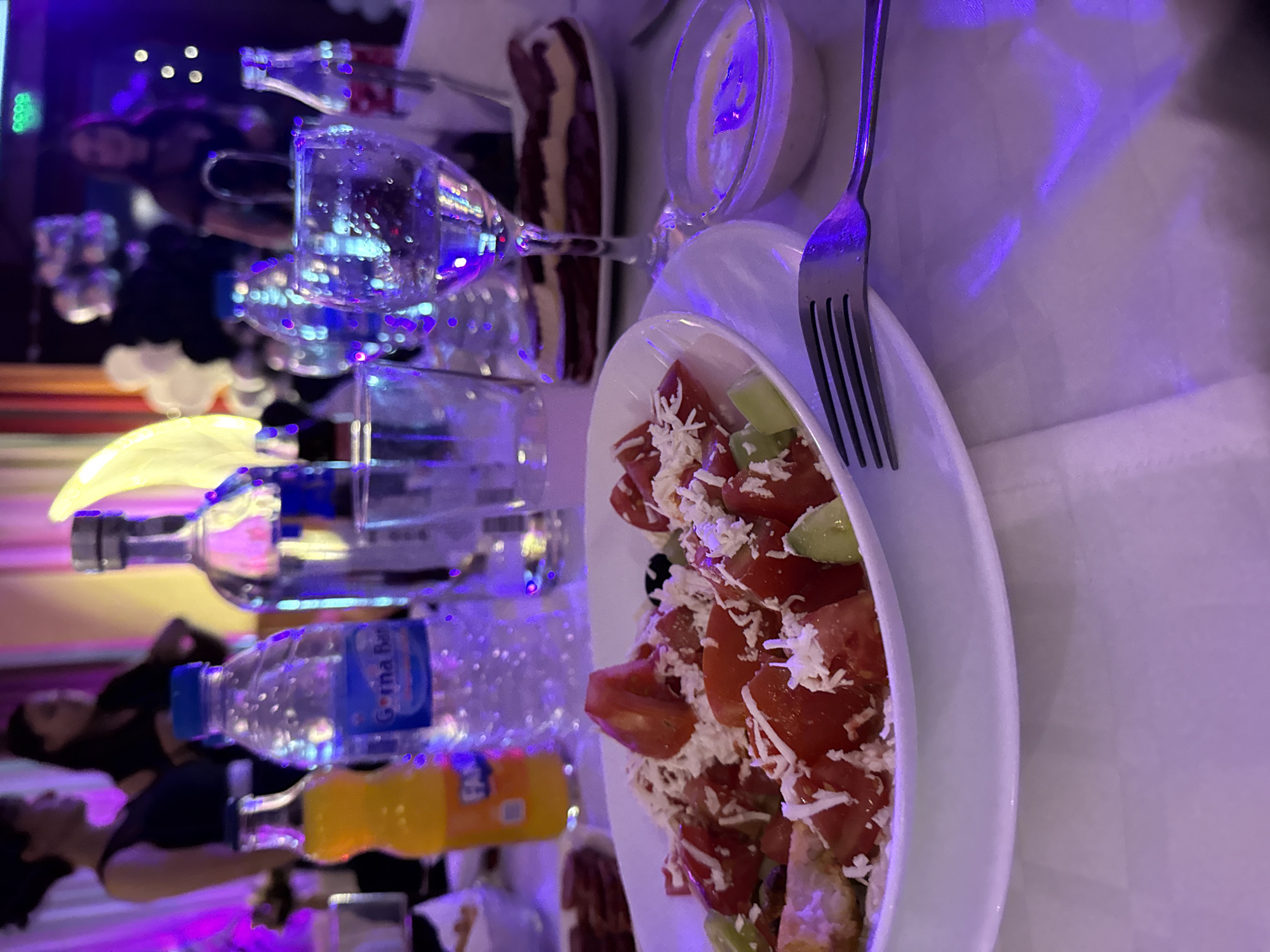 A table is set for a meal, possibly in a restaurant or event hall in Elhovo, Bulgaria. The table is covered with a white tablecloth. A salad is in a white bowl and a fork is on the right of the bowl. Beside the bowl is a small dish of white sauce. A row of glass water bottles and drink bottles, topped by multiple clear drinking glasses, are on the left side of the frame. There are several blurry human figures in the background. The lighting is dim and a purple/blue tone.