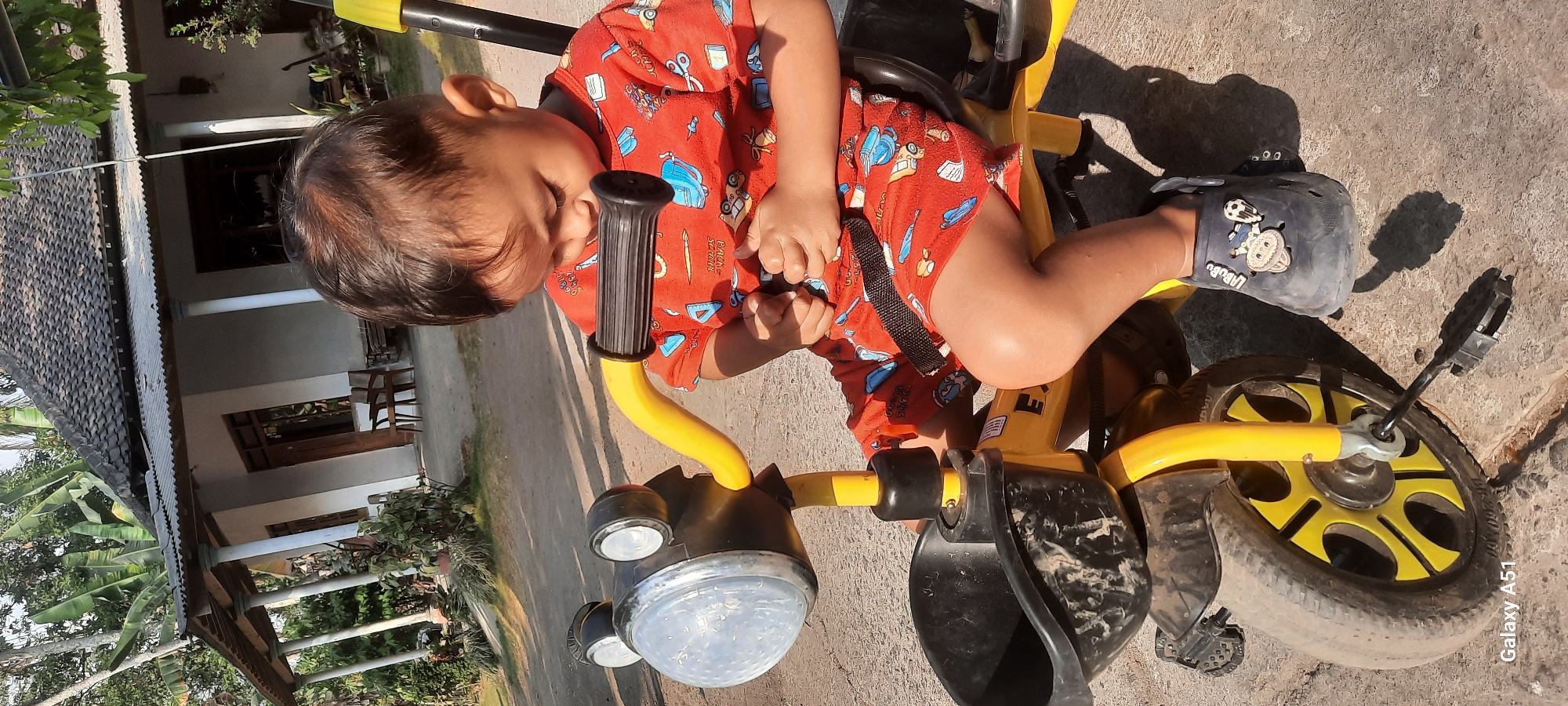 A young child, appearing to be of Indonesian descent, sits on a yellow tricycle in what appears to be a residential outdoor setting. The child is wearing a red t-shirt with a pattern of various cartoon objects and a pair of red shorts with a similar pattern. The child's legs are bent, and one foot, clad in a dark blue shoe, rests on the tricycle's pedal. The tricycle has a black basket in front and a yellow handlebar. Behind the tricycle, a house with a dark tiled roof and white walls is visible, with trees and vegetation surrounding it. The ground is paved with concrete or asphalt. The overall lighting suggests a daytime scene. The scene is consistent with a residential area in Ploso, Indonesia, given the architectural style of the house and the tropical vegetation.