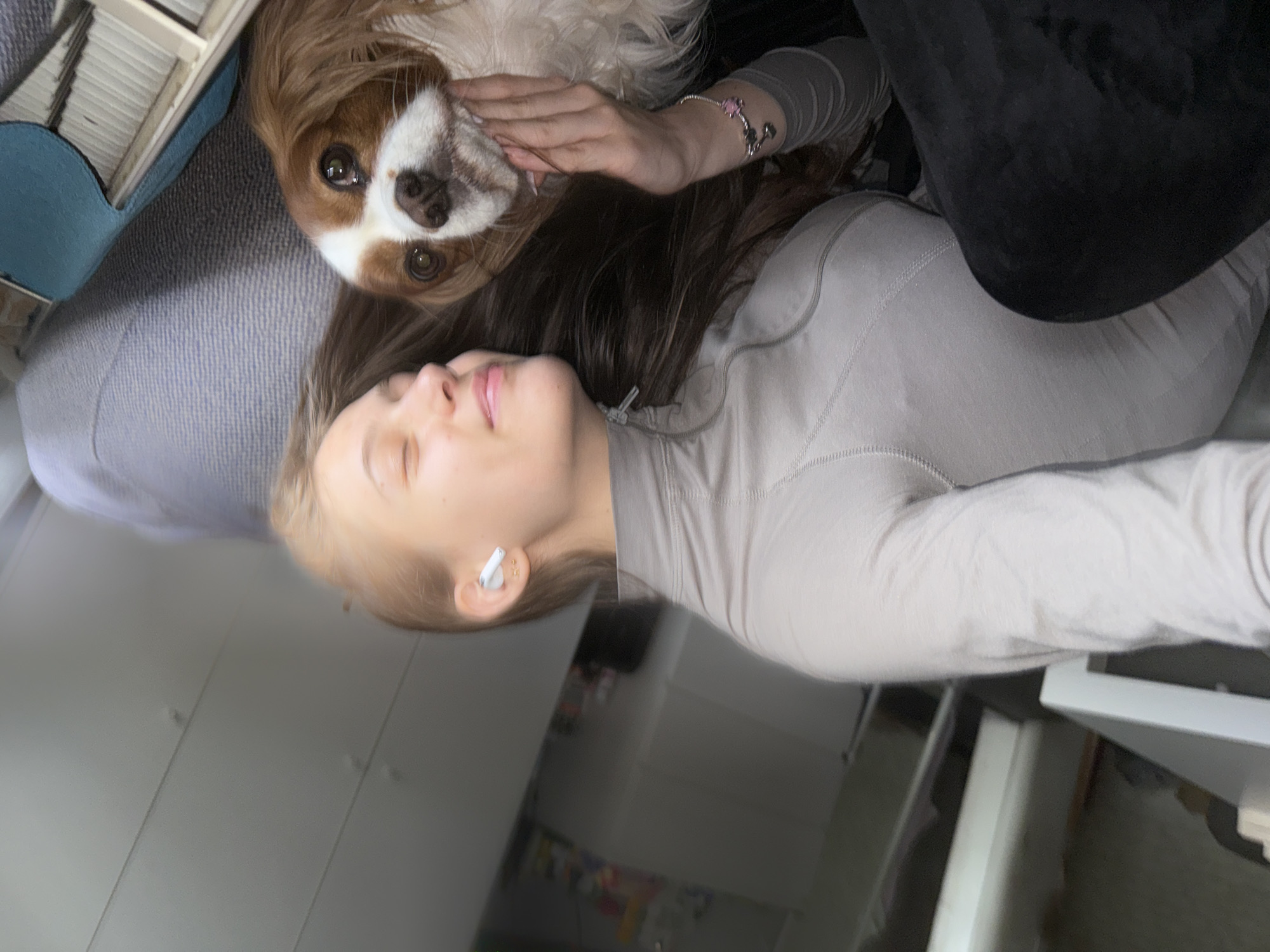 A Caucasian woman with fair skin and light blonde hair is reclining on a light gray textured surface, possibly a couch or bed. She is wearing a light gray turtleneck long-sleeved shirt and has a white earbud in her left ear. Her eyes are closed, and her expression is relaxed.
Lying next to her is a Cavalier King Charles Spaniel with a brown and white coat. A hand, presumably belonging to the woman, is resting on the dog's face.
The background is blurred but appears to show a room with white furniture. The location in Sofia, Bulgaria is not discernible from the image.
