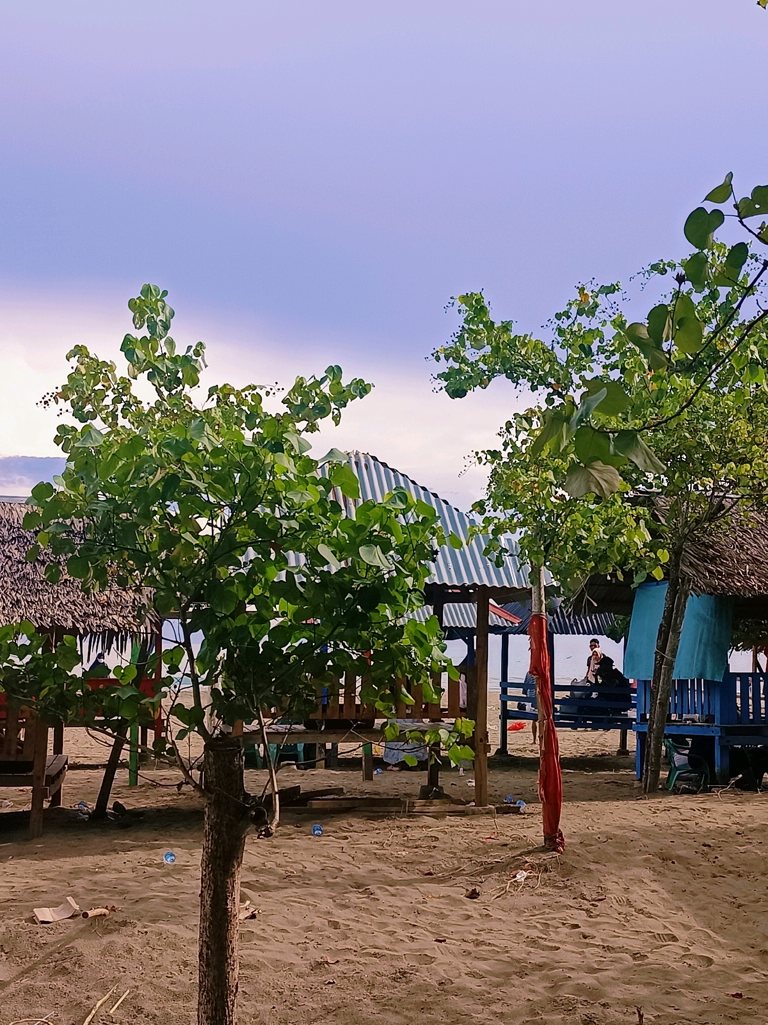 The image depicts a serene beach scene in Reudeuep, Indonesia. In the foreground, a young tree with lush green leaves stands prominently on the sandy shore. Beyond it, a row of simple thatched-roof huts and covered seating areas line the beach, offering shade. The huts feature striped canvas roofs and some have blue wooden furniture. Two people are visible in the background, one seated and another standing, near the water's edge. The sand is lightly textured with footprints and scattered debris, suggesting recent activity. The sky above is a soft, hazy blue with hints of clouds. The overall atmosphere is tranquil, capturing a typical day at this Indonesian coastal location.
