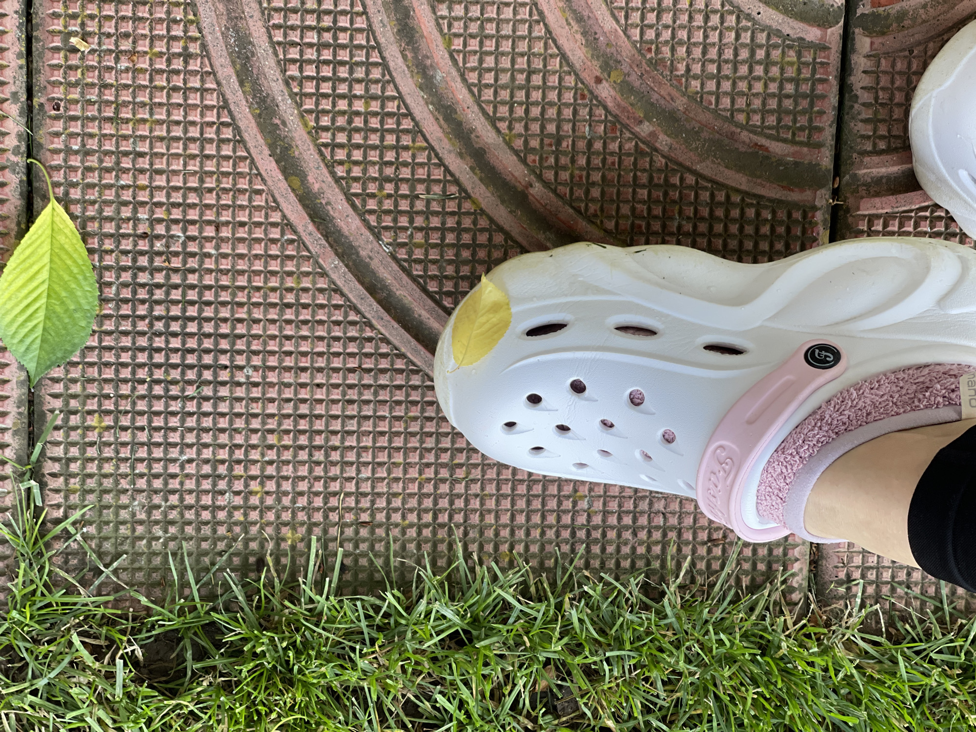 A close-up photograph shows a person wearing a white Croc-style clog with a pink strap and fluffy interior trim, standing on a reddish-brown tiled surface with a geometric pattern. The shoe is on the right side of the frame, partially cropped, with the person's foot and black-clad leg visible inside. To the left of the shoe, a bright green leaf rests on the tiles. Along the bottom edge of the photo, green grass is visible. The tiles appear to be part of a walkway or patio. Based on the type of tile and general aesthetic, the location is likely in an outdoor space in Sofia, Bulgaria, but there are no specific landmarks visible to definitively confirm this. The image suggests a casual, outdoor setting.