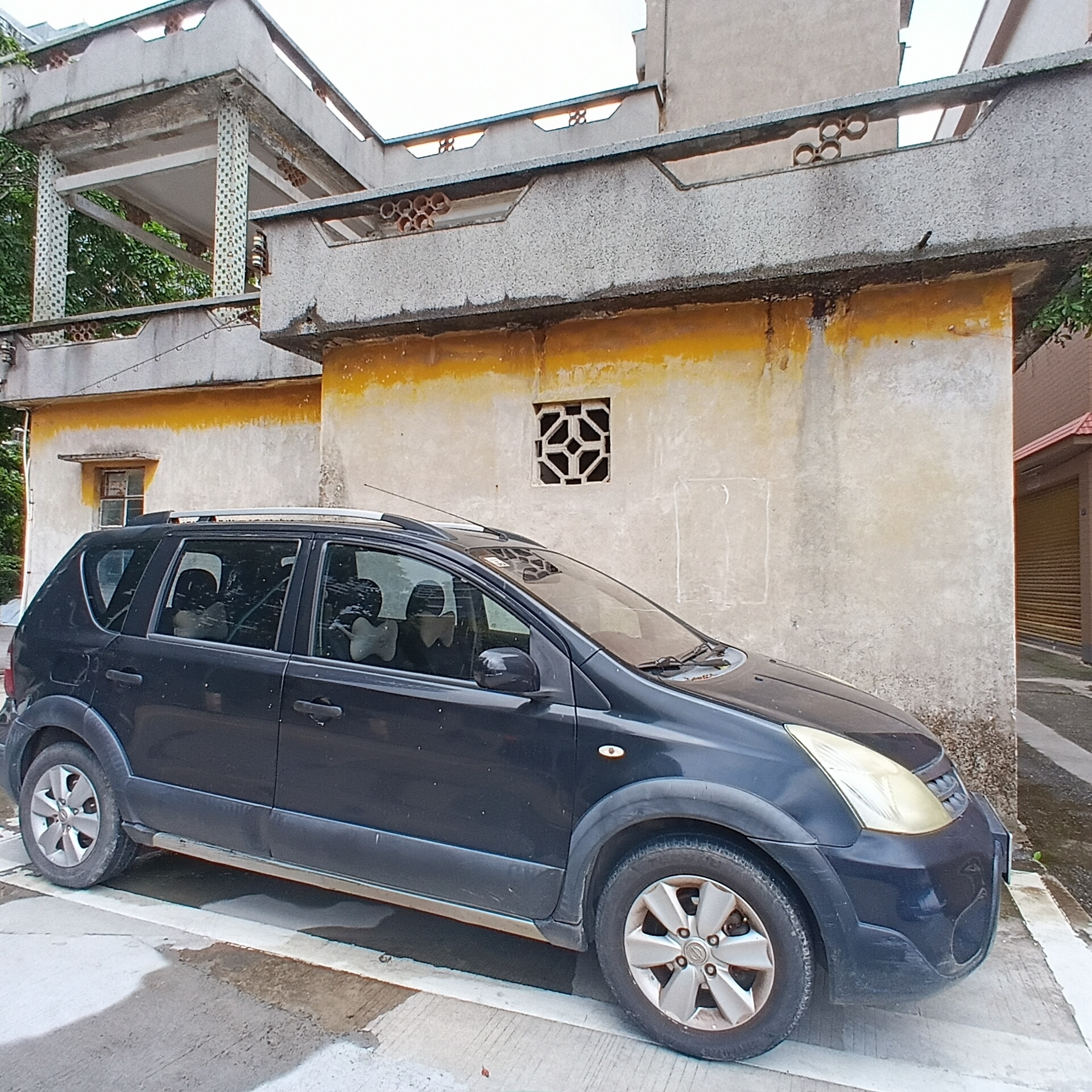 A black Nissan Grand Livina compact MPV is parked in front of a two-story, concrete building. The building has a yellow horizontal line along the top of the first story. The upper story is a covered porch. The wall to the right of the vehicle has a square decorative opening. The scene is in the Shunde District, China.