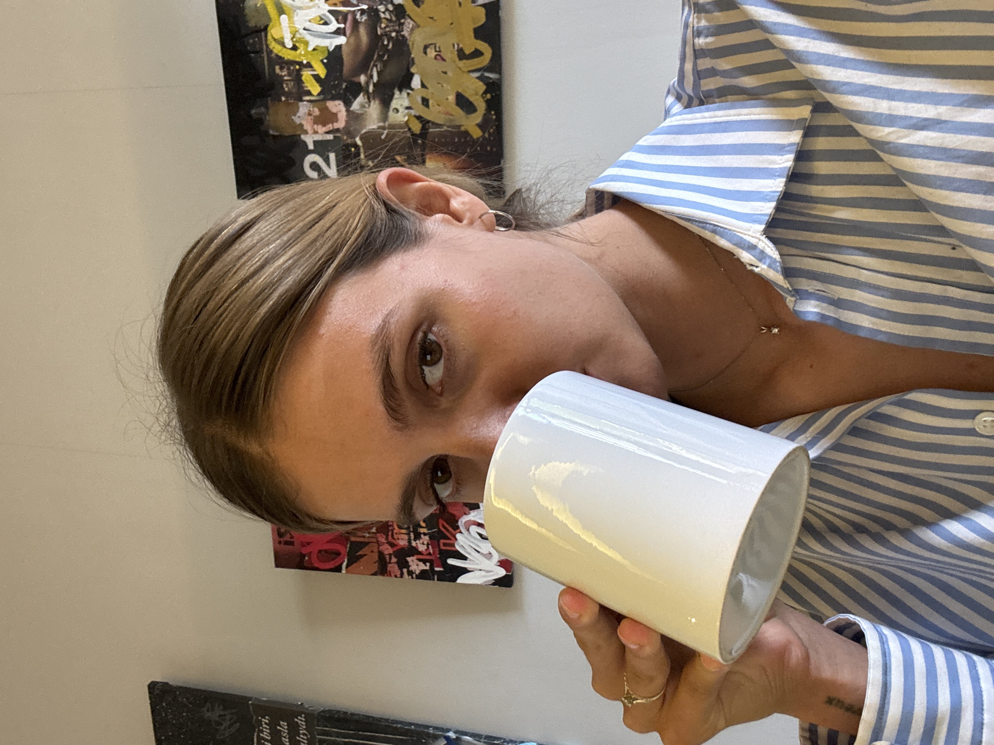 A close-up, portrait-oriented image, rotated 90 degrees counter-clockwise, depicts a person, likely female, holding a white cylindrical mug or tumbler. The person has light brown hair, pulled back, and is wearing a blue and white striped collared shirt. They are looking directly at the viewer with their right brown eye, while the left eye is obscured by the object they hold to their face.
Notable details include a small silver hoop earring in their visible ear. A delicate, silver-colored necklace with a small, star-shaped pendant is visible on their chest. On their right ring finger, a ring with a star-shaped design is worn. A small, dark tattoo, possibly displaying Roman numerals or a date, can be seen on their inner right wrist.
The background features a plain, light-colored wall adorned with several art pieces. In the upper section of the frame, there is an abstract artwork characterized by yellow, black, and white elements, with the number "210" clearly visible. Below and to the left, another art piece with red, black, and white graffiti-style markings is partially shown. In the lower left, a dark book or framed item displays text in a foreign language, with words such as "birî,", "asla," and "ralyılı" partially legible. The lighting is bright, suggesting an indoor environment.