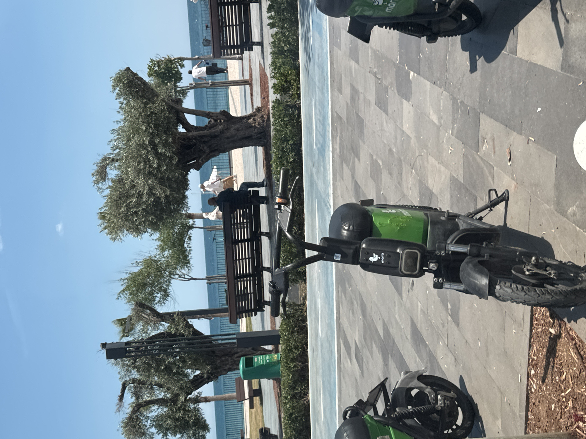 The scene depicts a seaside walkway in Konyaaltı, Turkey, identifiable by the Martı brand shared electric scooters in the foreground. The path is paved with grey rectangular stones alongside a narrow strip of blue and green surface that separates the walkway from a line of trees and benches. Two people are seated on a bench under a gnarled, mature tree. Additional figures can be seen walking further along the path towards the sea. The sea is visible in the background, with a line of buildings or structures along the distant coast. The sky is clear and blue.